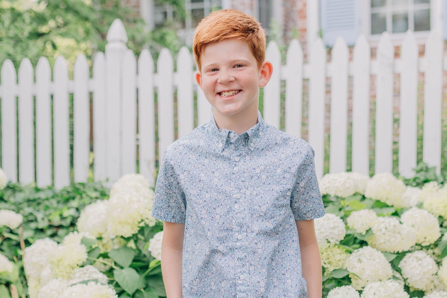 Smiling red-haired boy in a blue floral shirt standing in front of white hydrangeas and a picket fence.