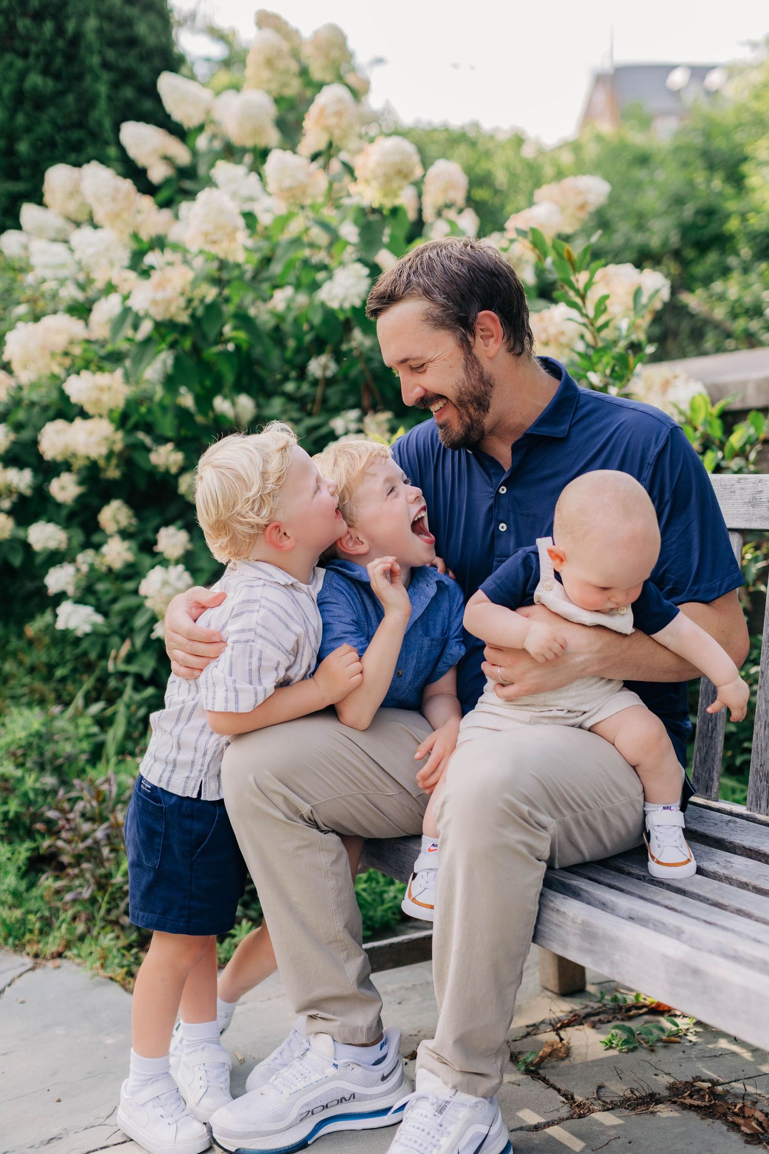 A father sits on a garden bench holding a baby while two young boys laugh and play around him near white hydrangeas.