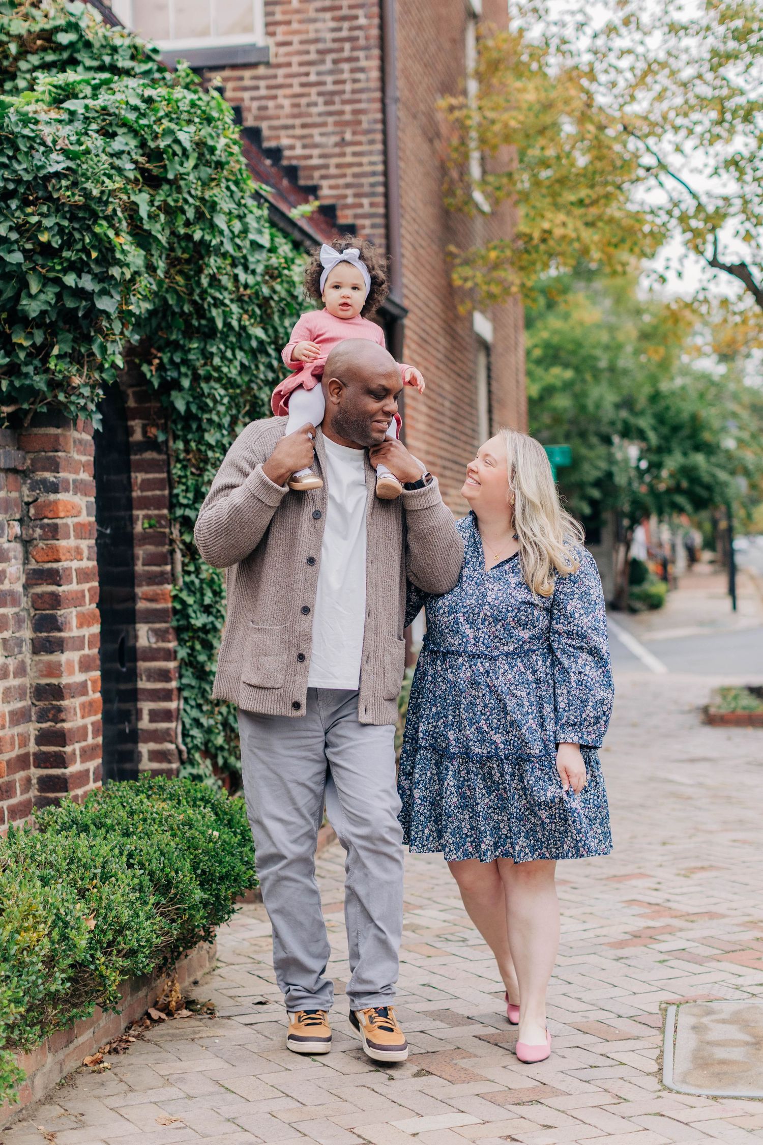 A happy interracial family strolling on a brick sidewalk, with dad carrying baby on shoulders and mom in floral dress.