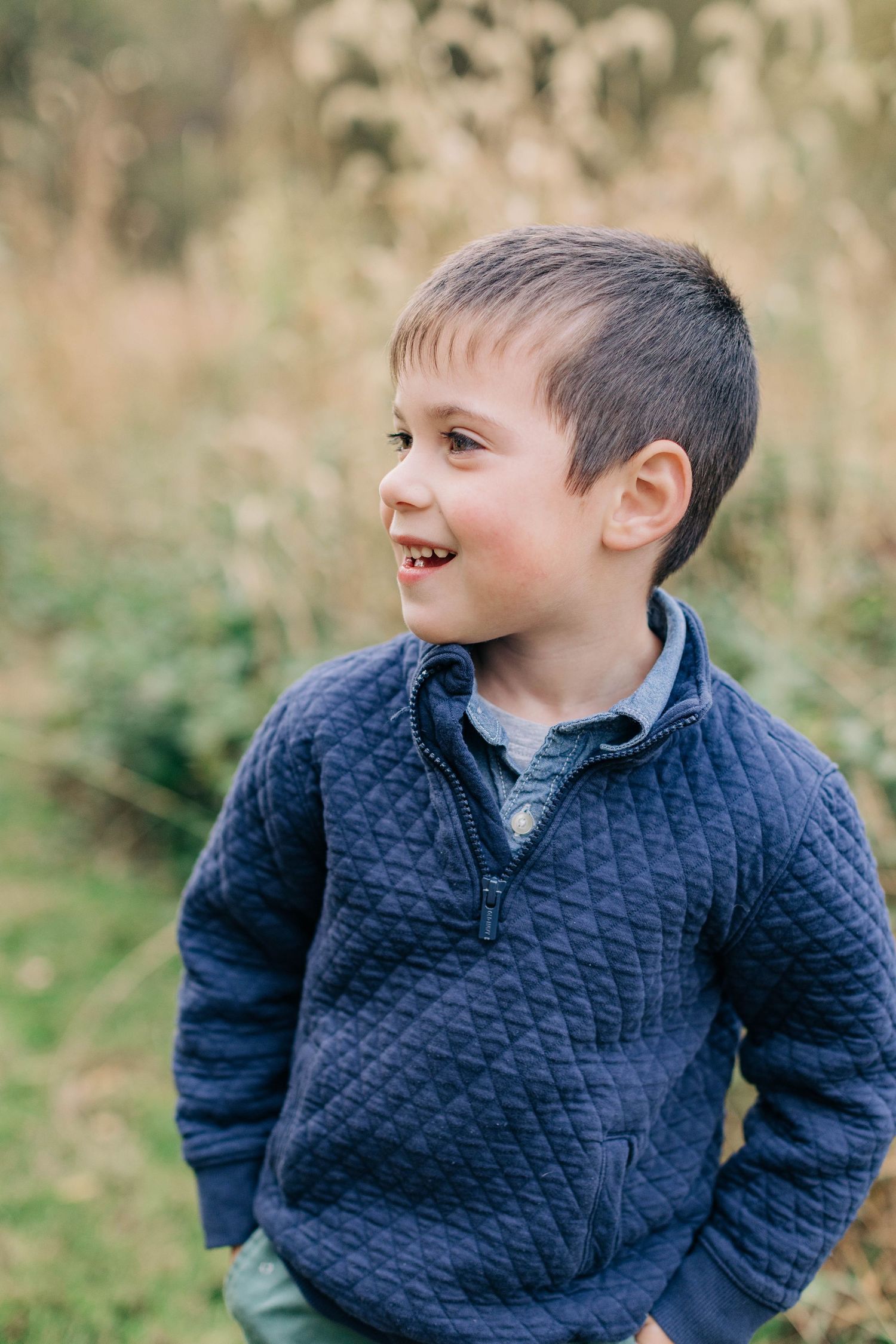 Young boy in navy quilted pullover smiling outdoors in autumn field with golden grasses in background.