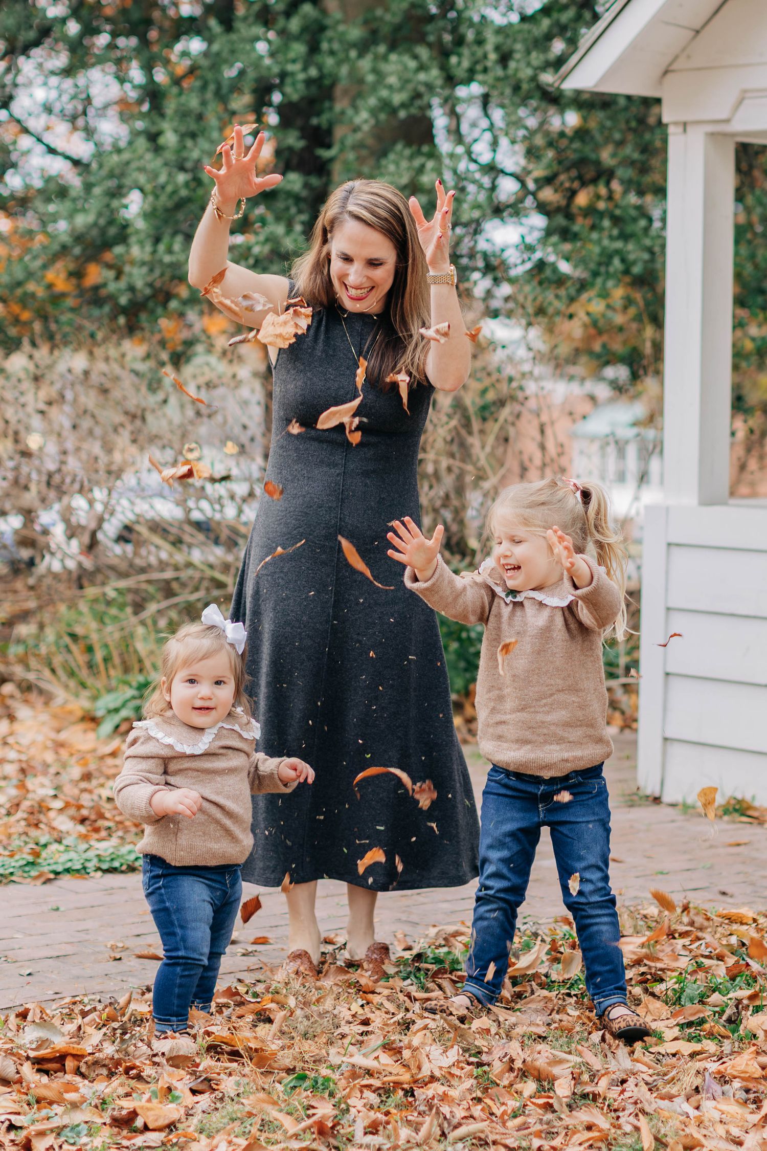 Mother and two young daughters joyfully tossing autumn leaves outdoors in matching gray and tan outfits.