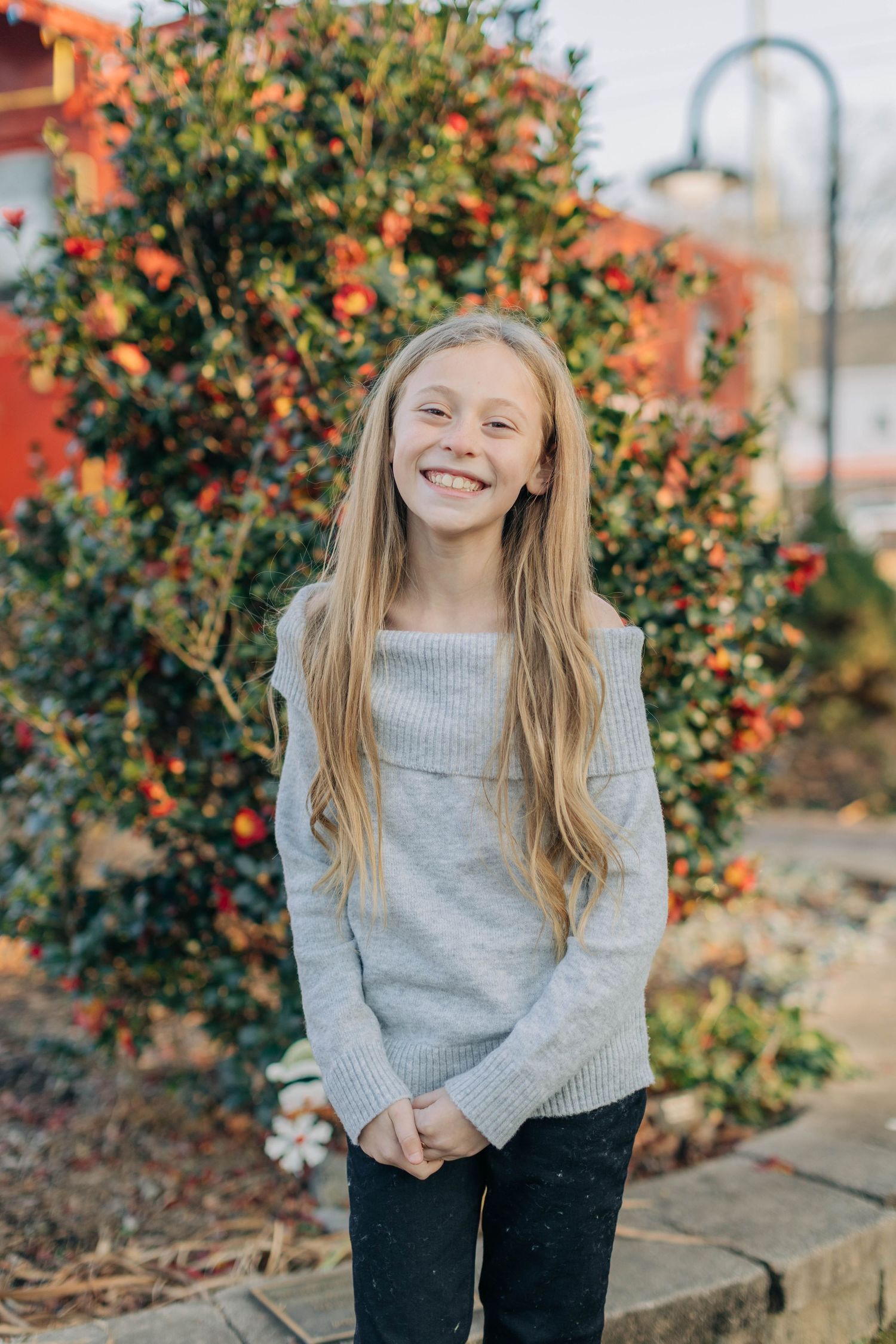 Smiling young girl with long blonde hair wearing a gray sweater standing outdoors near a berry bush.