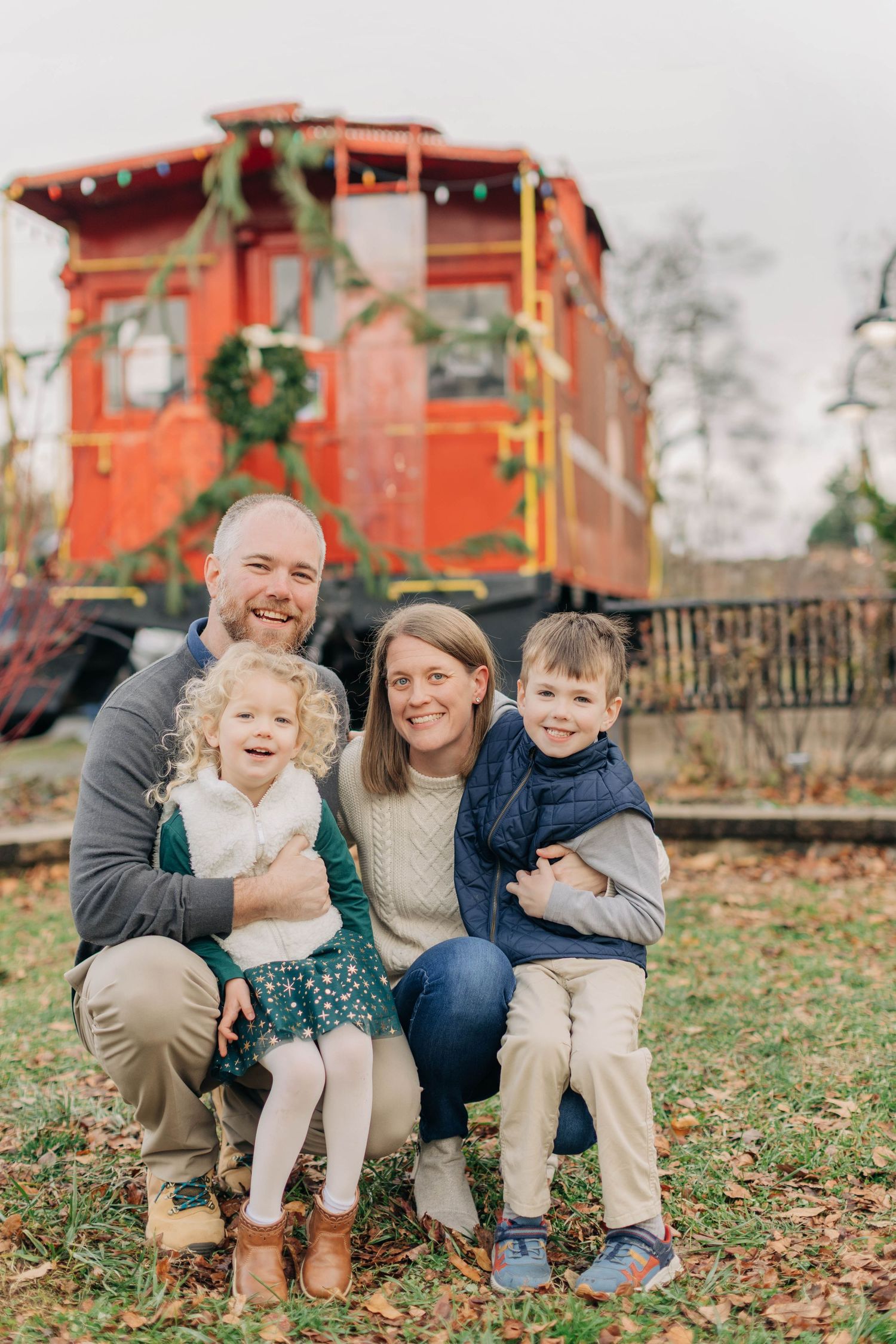 Family of four smiling in front of a decorated red train car with holiday wreaths during a festive outdoor photo session.
