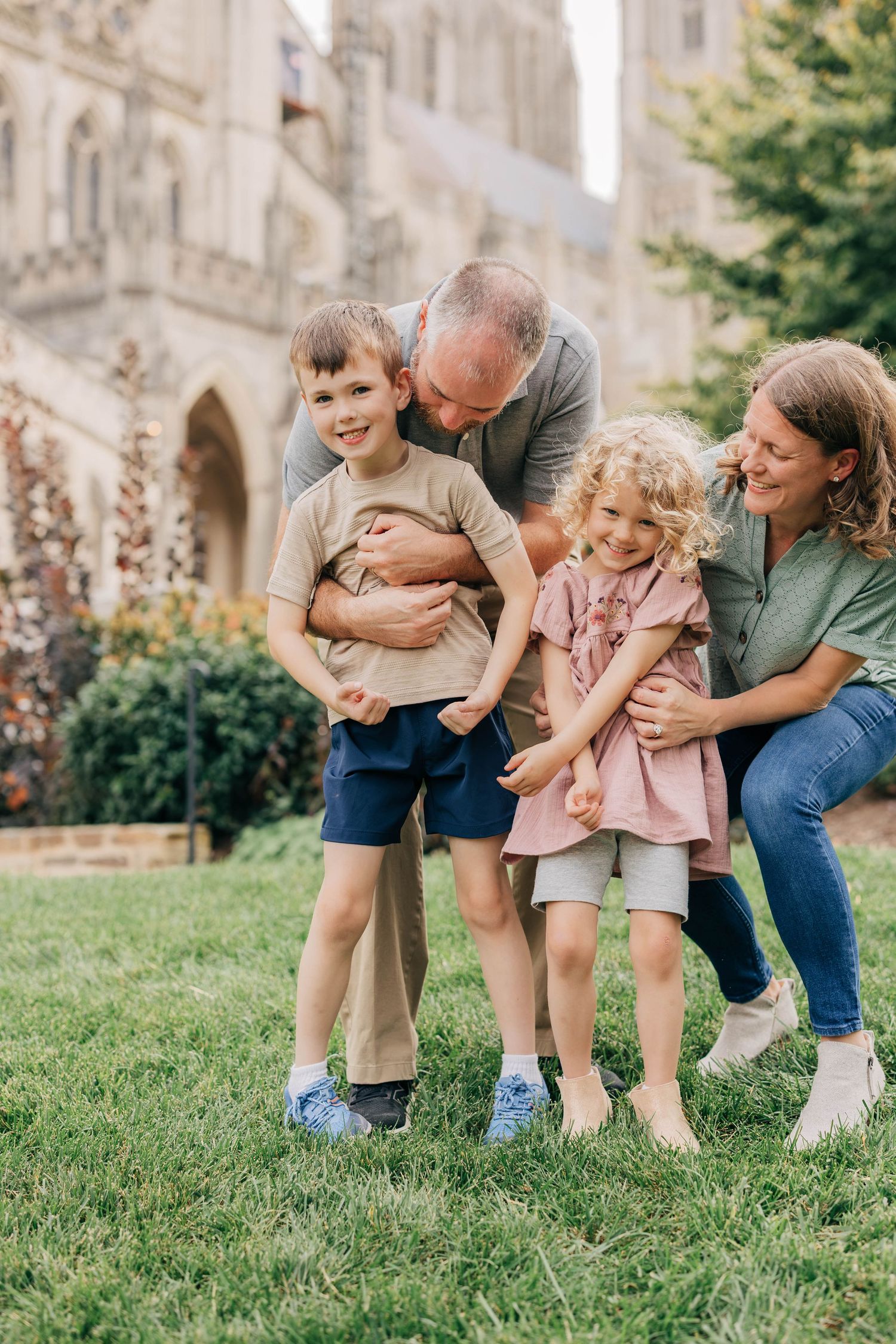 Smiling family of four hugging and laughing on a sunny lawn in front of a historic Gothic stone cathedral.