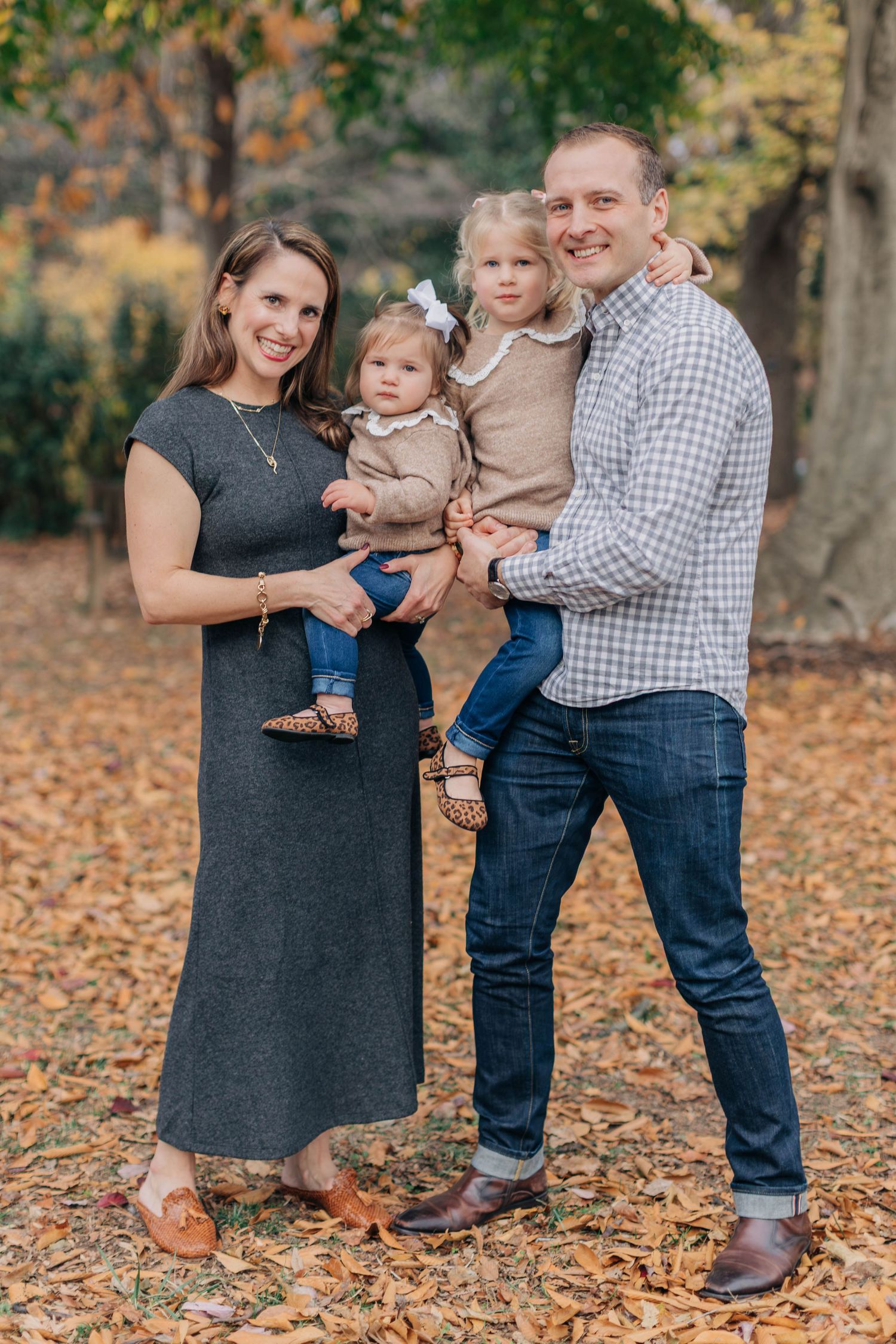 Family of four posing outdoors in fall foliage, parents holding two young daughters in coordinating neutral outfits.