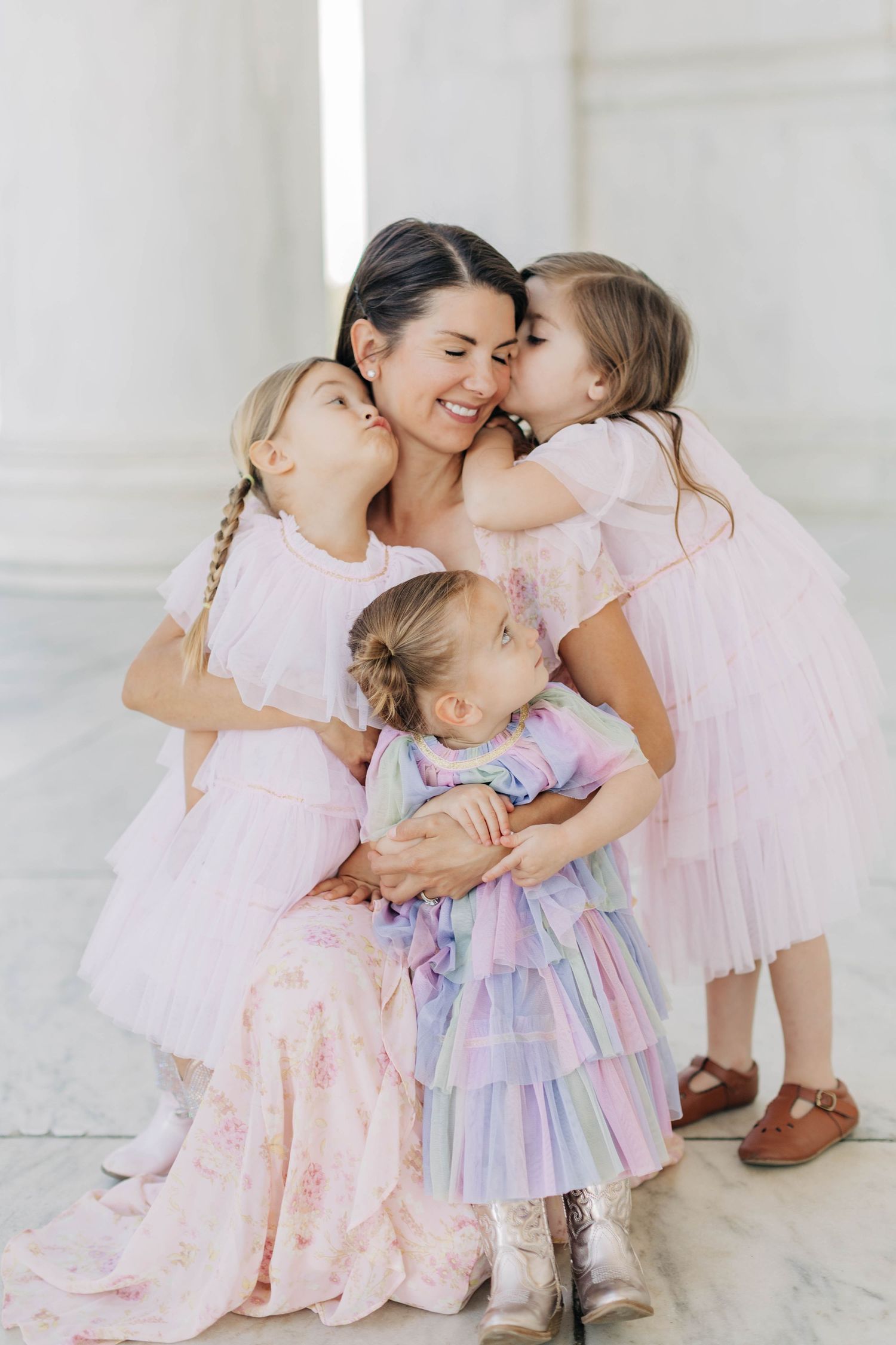 A mother kneels as three young daughters in pink tulle dresses embrace her with hugs and kisses.