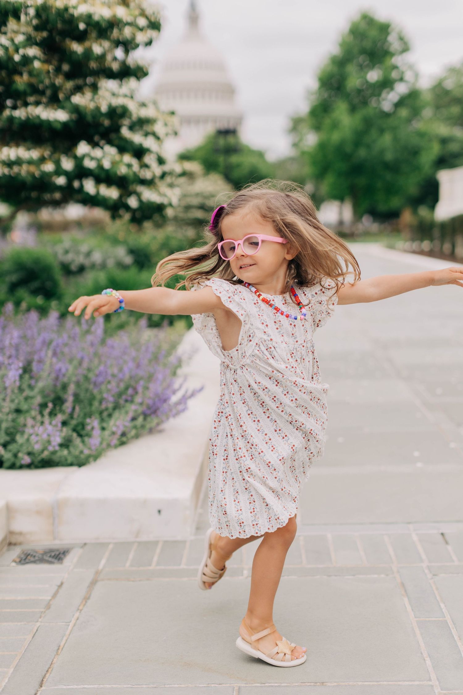 A joyful little girl in a floral dress and pink glasses twirls on a stone path near the US Capitol building.