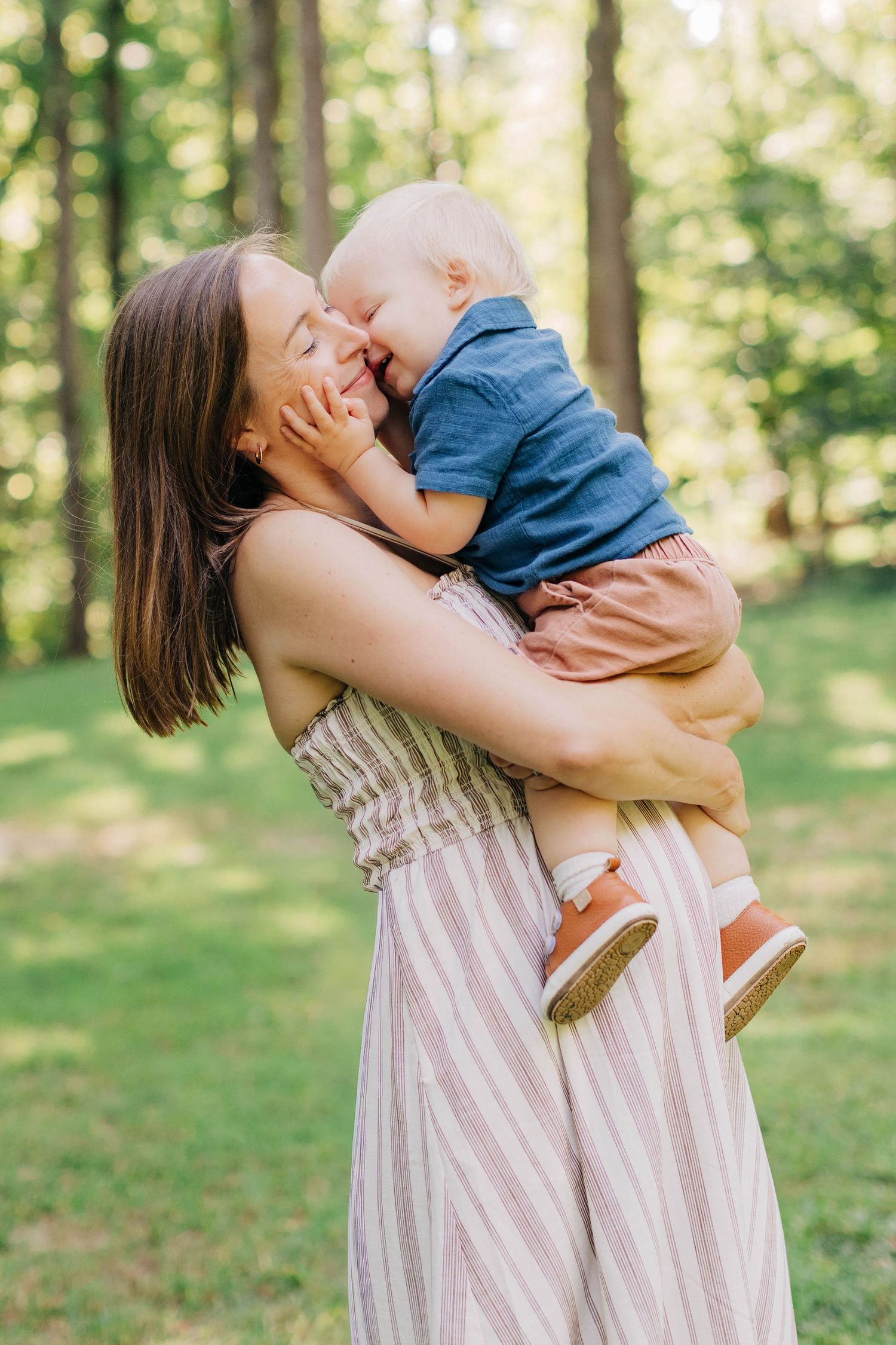 A young mother in a striped dress holds and nuzzles her baby boy outdoors in a lush green park.
