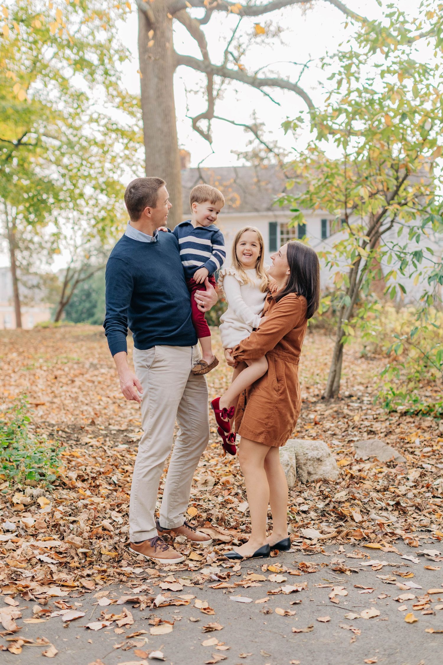 Family of four enjoying autumn outdoors, parents holding two young children among colorful fallen leaves near a historic home.