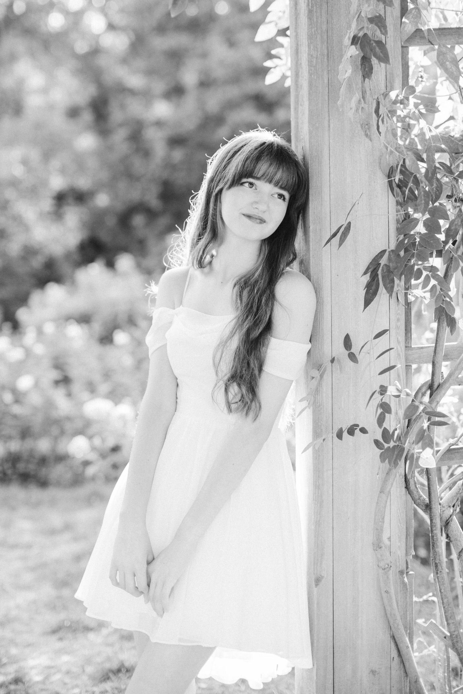 Ethereal black and white portrait of someone in a white dress standing by a garden trellis.
