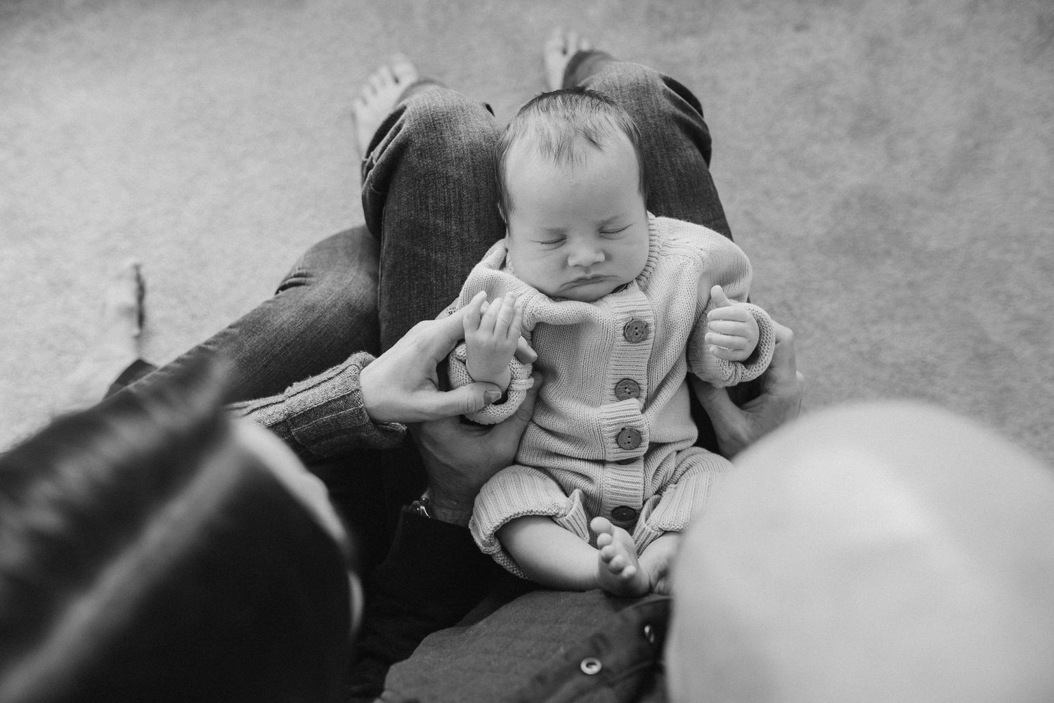 Black and white photo of baby in buttoned outfit sitting on cushioned surface.