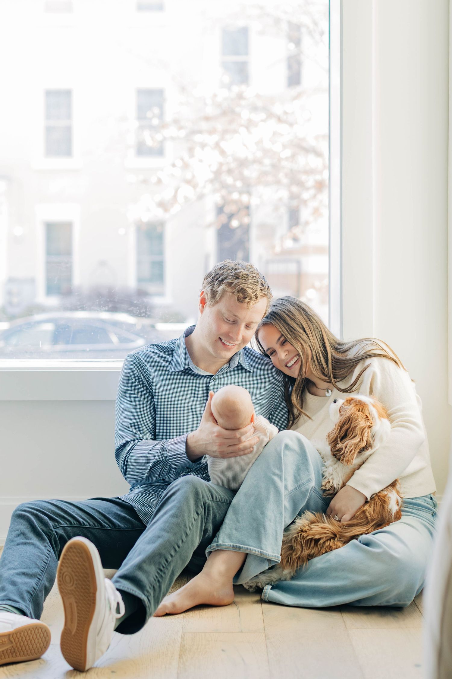 Couple sitting on floor by window holding newborn baby with golden spaniel dog in bright airy home.