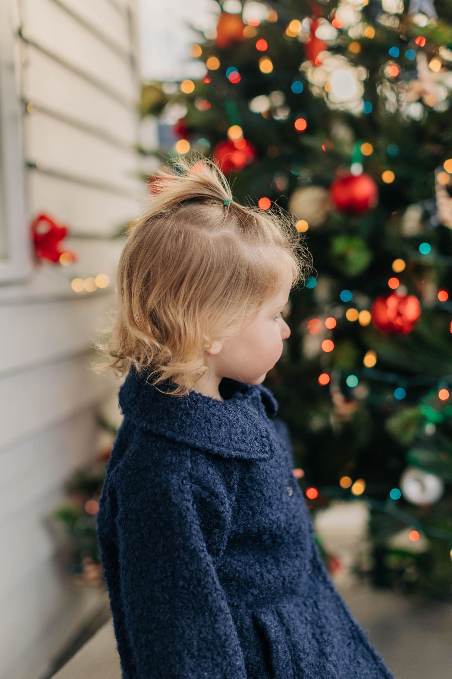 Toddler girl in navy coat standing near decorated Christmas tree with colorful lights and red ornaments.