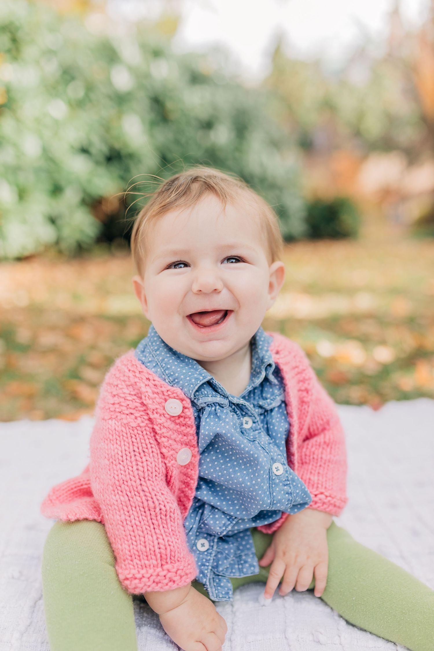 Smiling baby girl in pink knit cardigan and blue polka dot shirt sitting outdoors on autumn day.