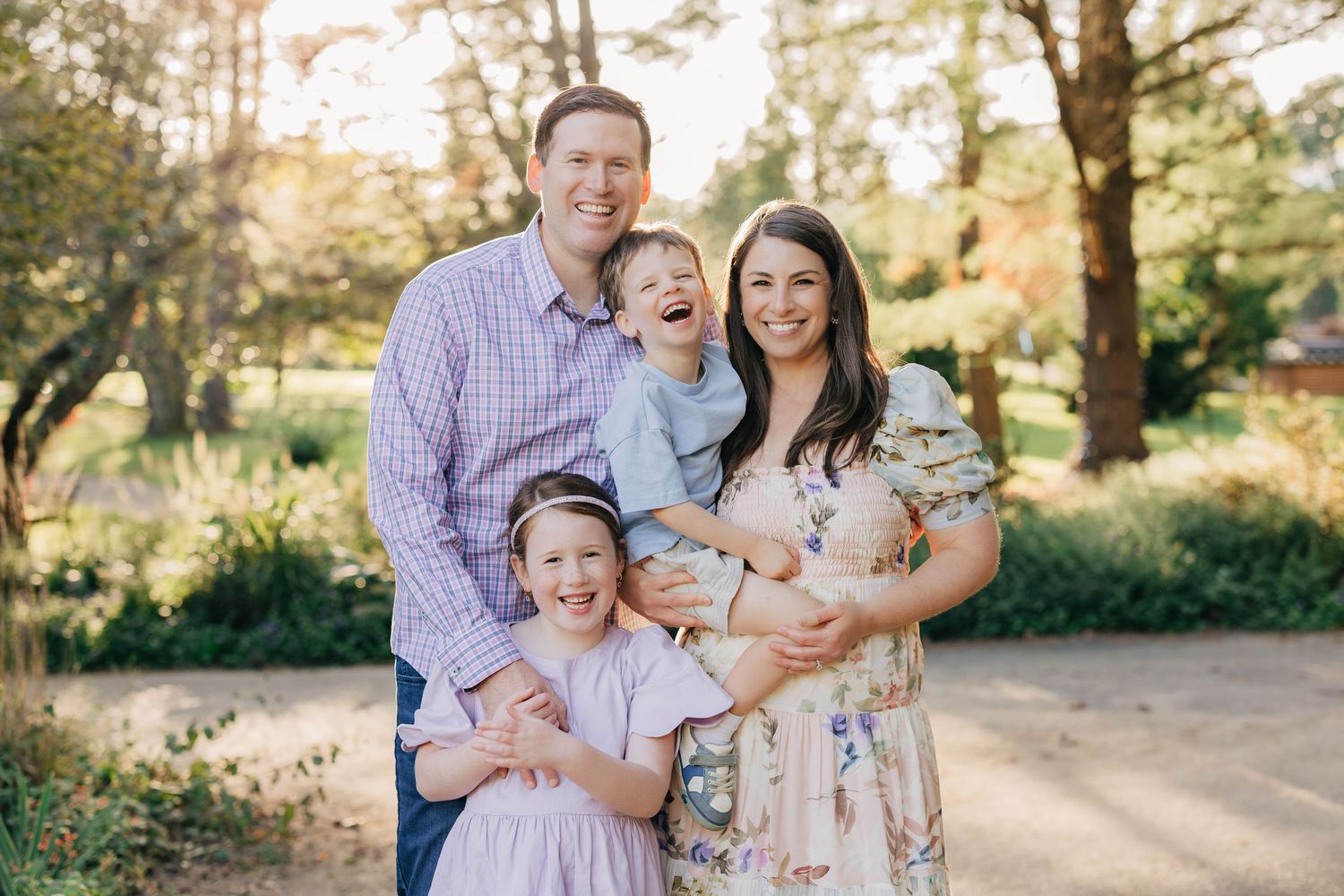 Happy family of five posing outdoors in a sunlit park, parents smiling with three young children in spring attire.