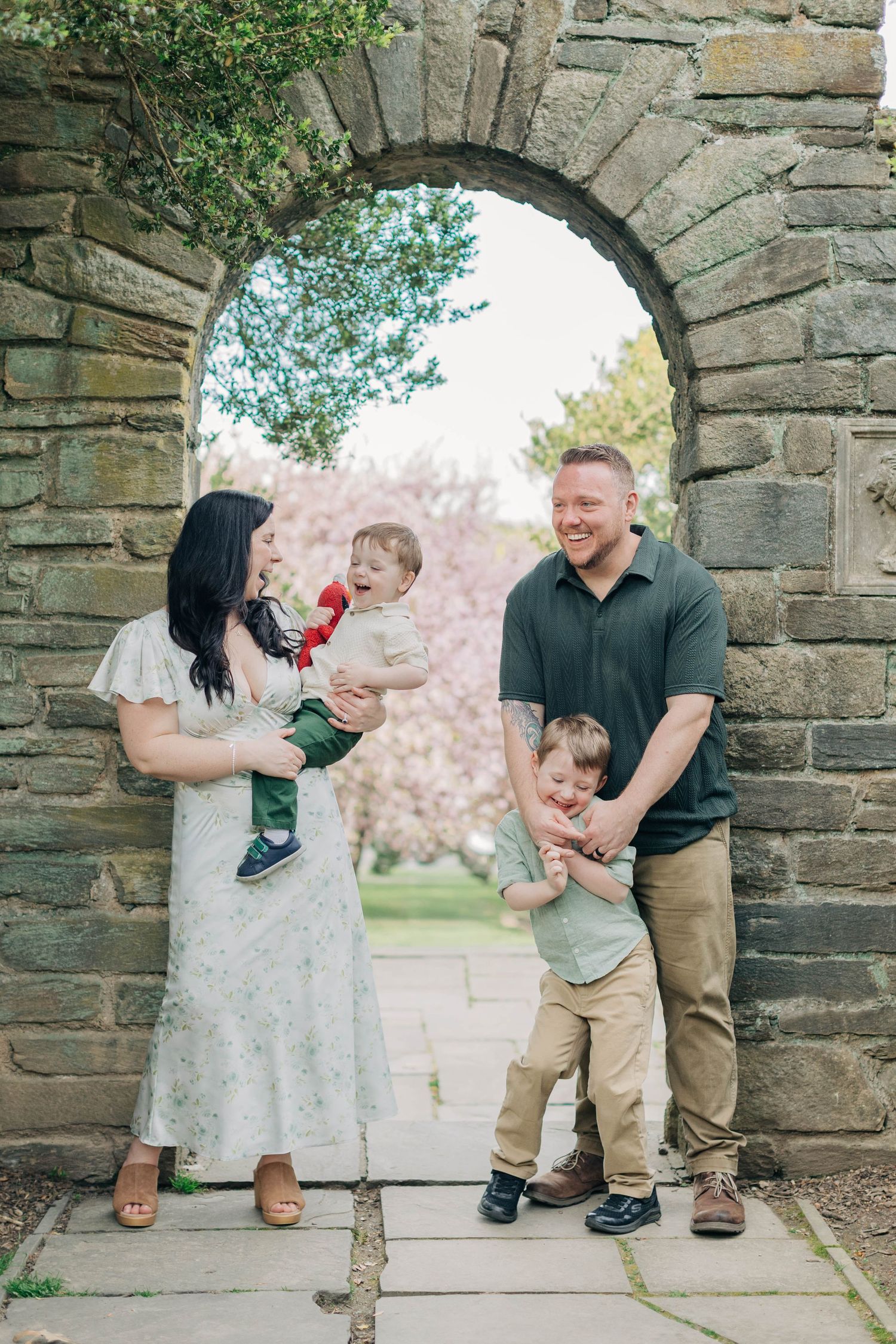 Family of four posing under stone arch with cherry blossoms, parents holding young children, smiling outdoors.