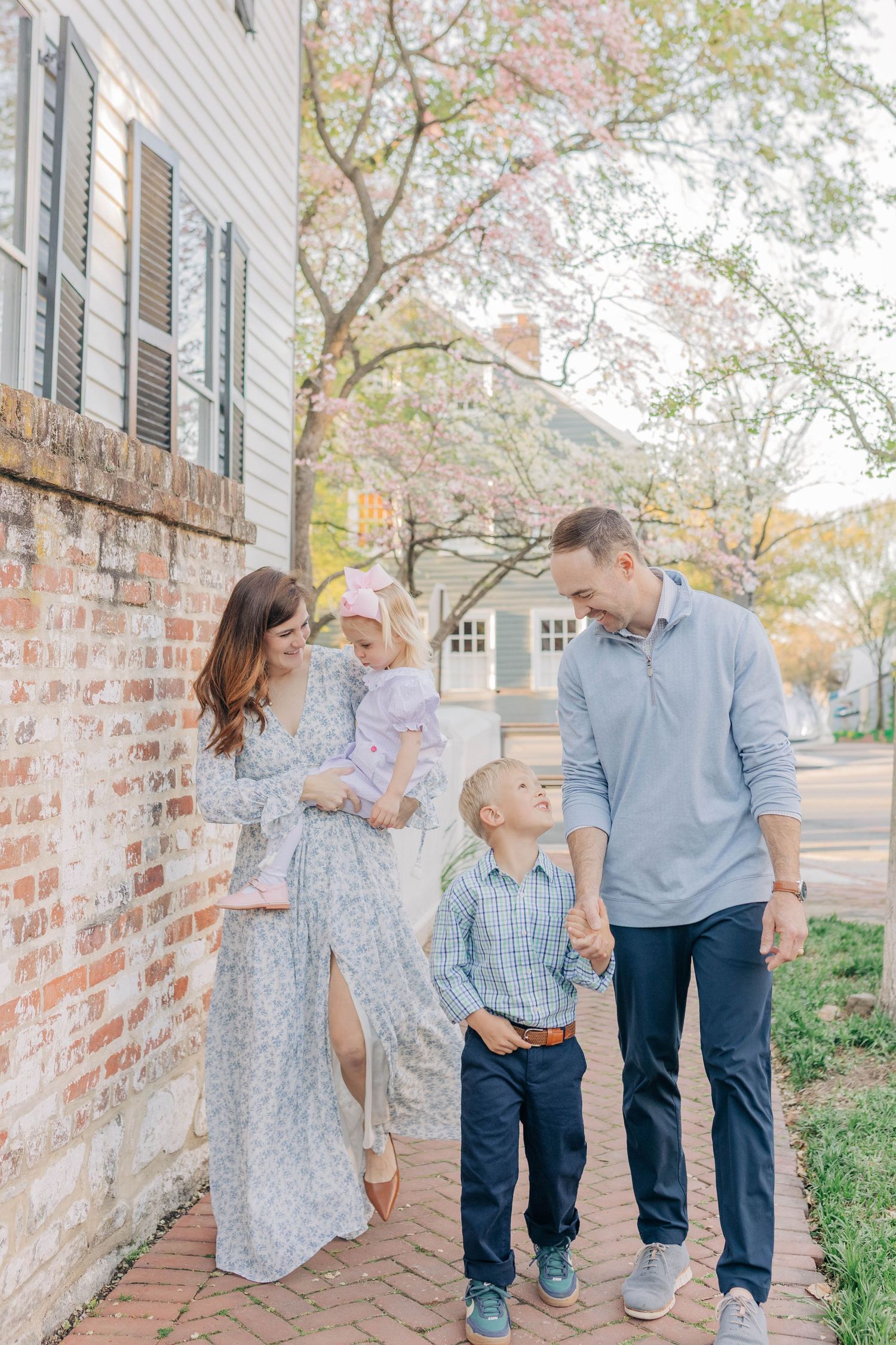 Family of four walking along a brick sidewalk in spring, mom holding toddler girl, dad holding young son's hand.