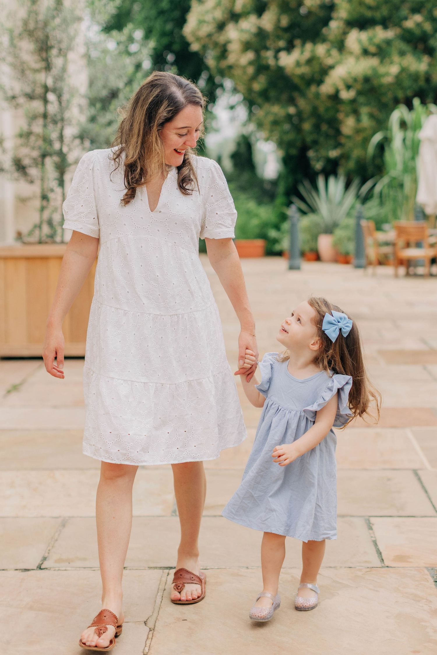 A mother in a white dress holds hands with her young daughter in a blue dress while walking outdoors.
