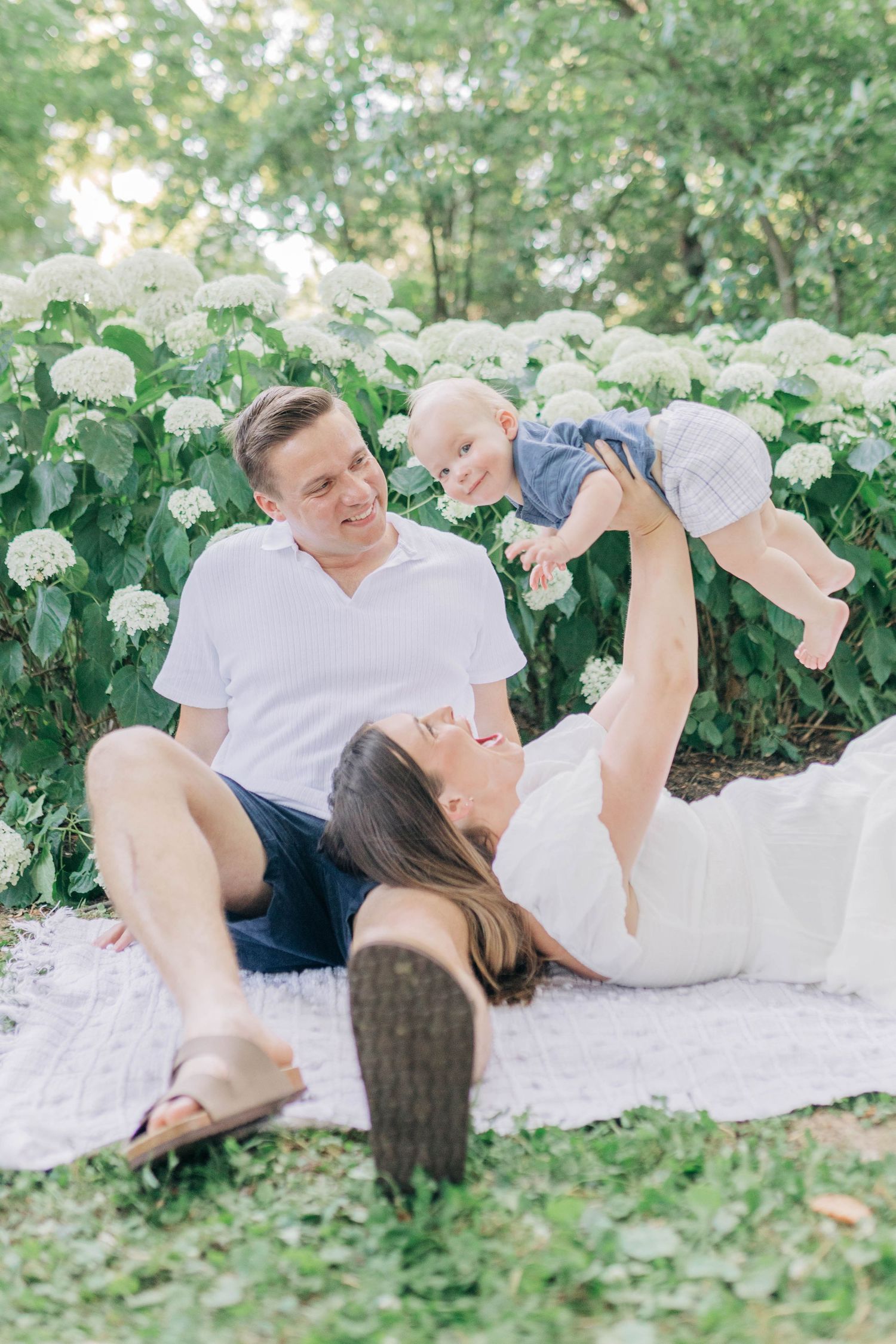 Family relaxes on white blanket in garden with blooming white flowers.