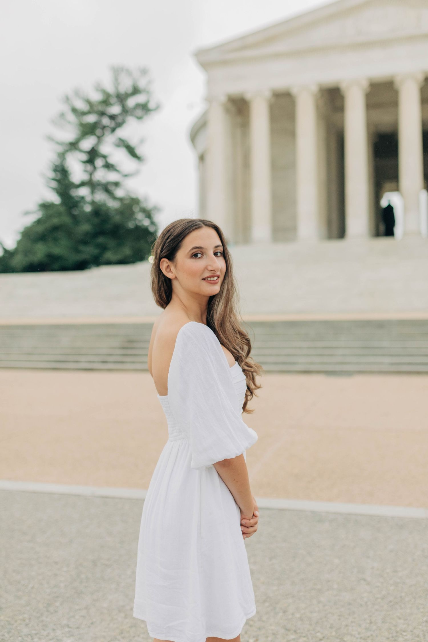 Person in white dress poses near a classical building with columns on a cloudy day.