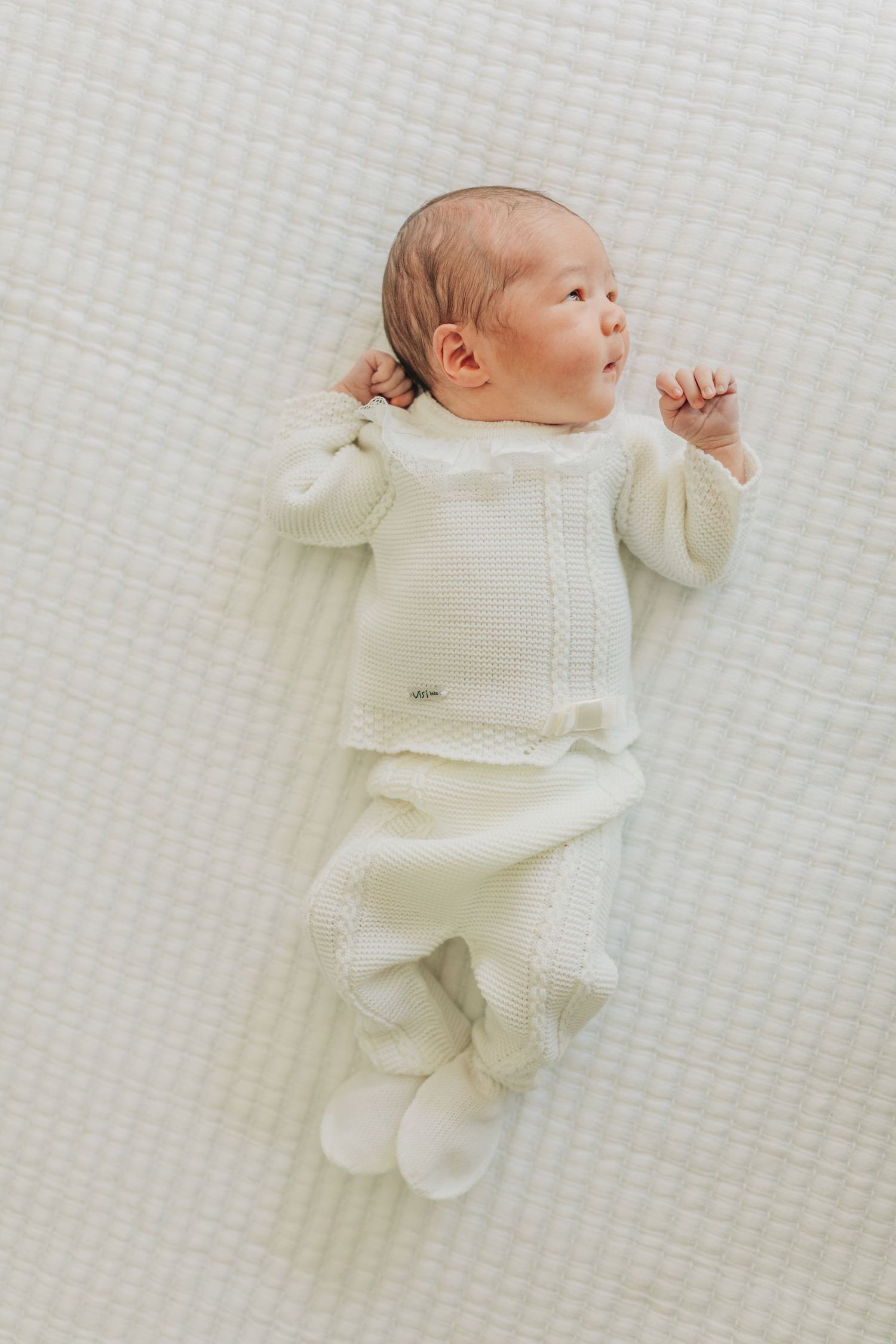 Baby wearing cream knit outfit lying on textured white surface.