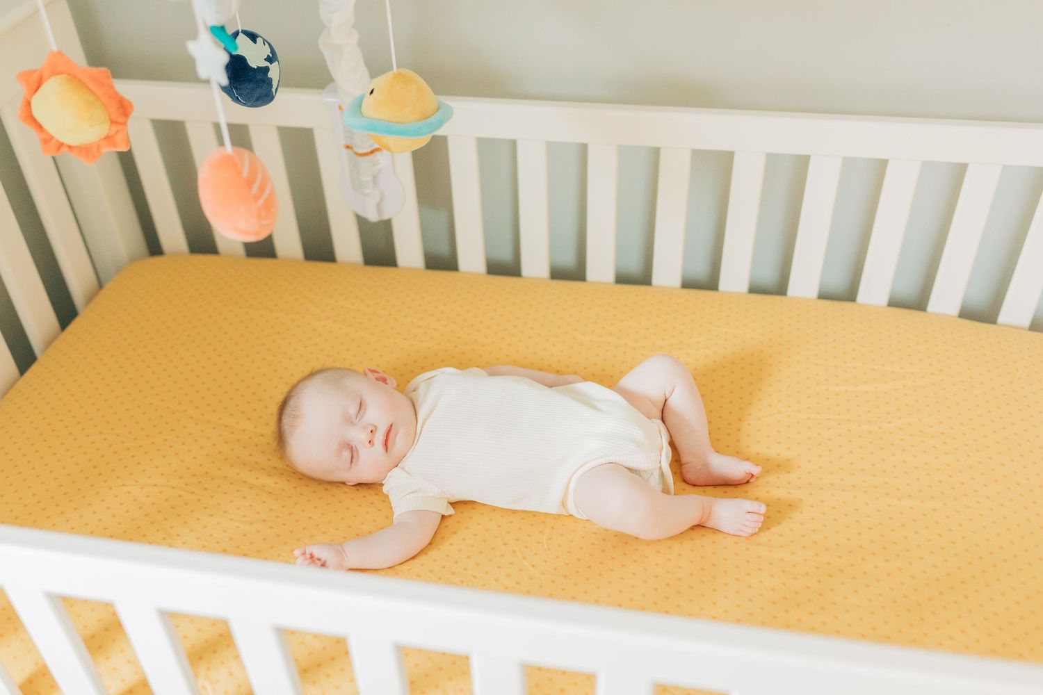 Infant resting on yellow crib mattress in nursery with white walls.
