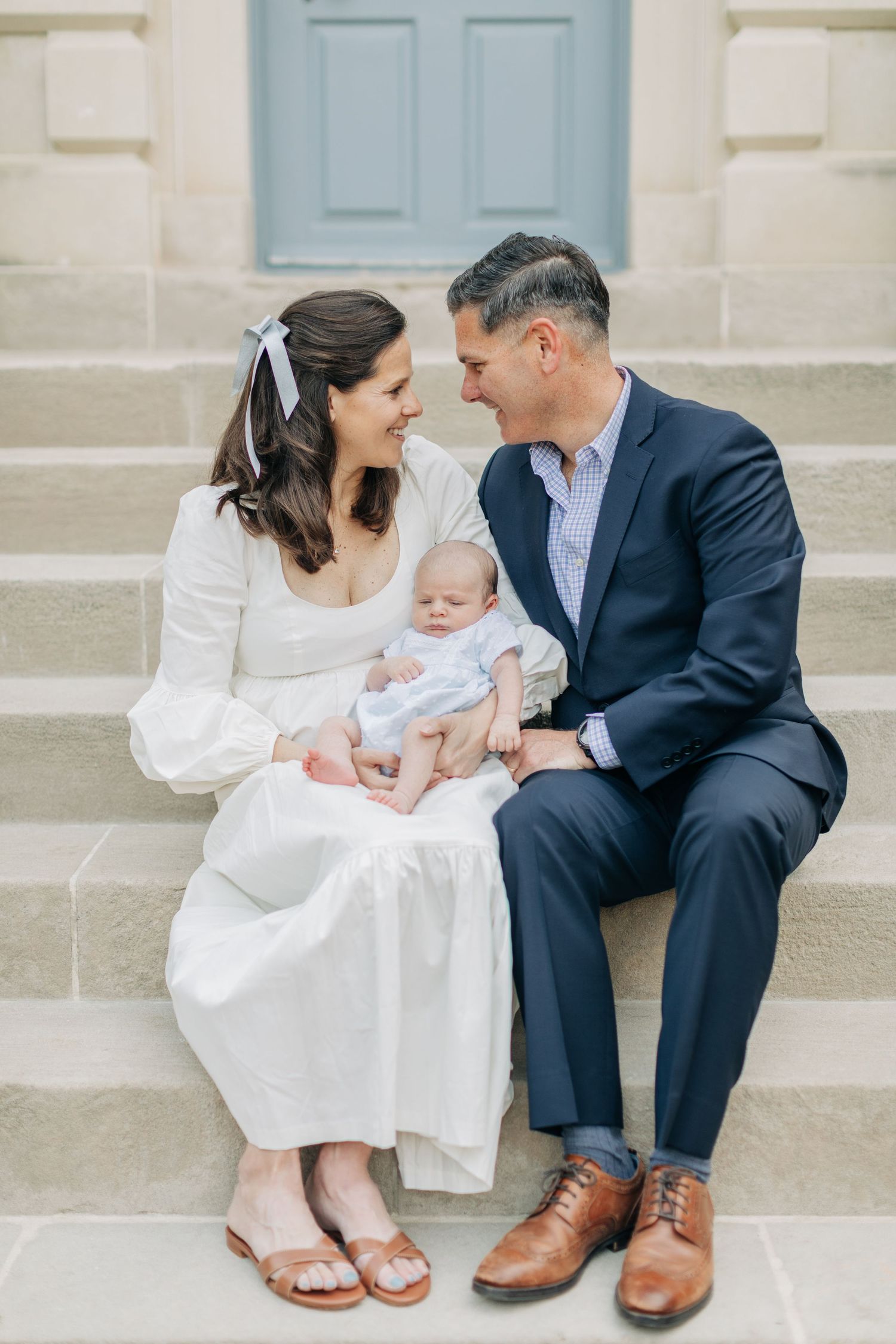 A couple sits on stone steps with their baby wearing white and navy formal attire.