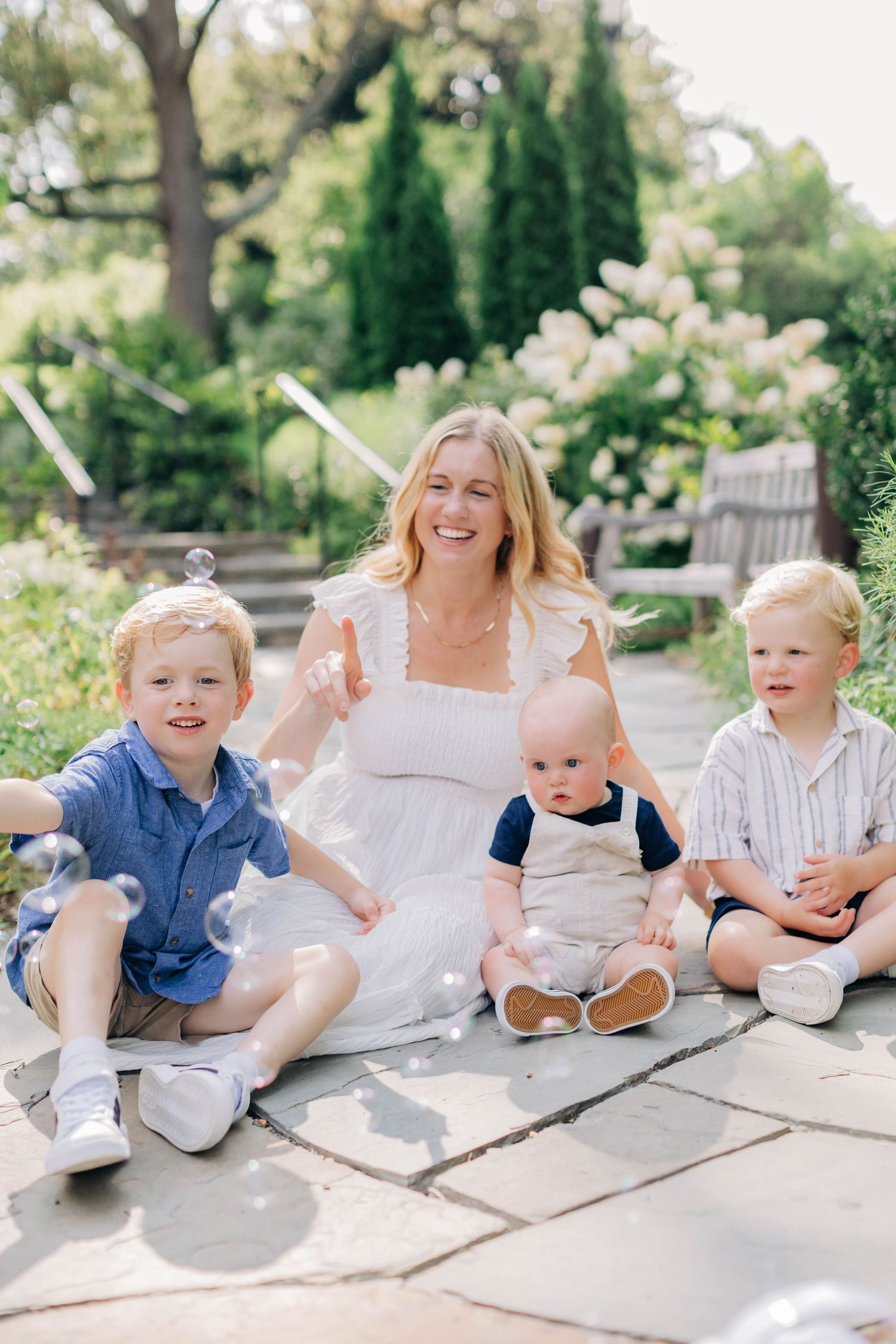 A smiling blonde woman in white dress sits on stone path with three young boys and bubbles in a lush garden.