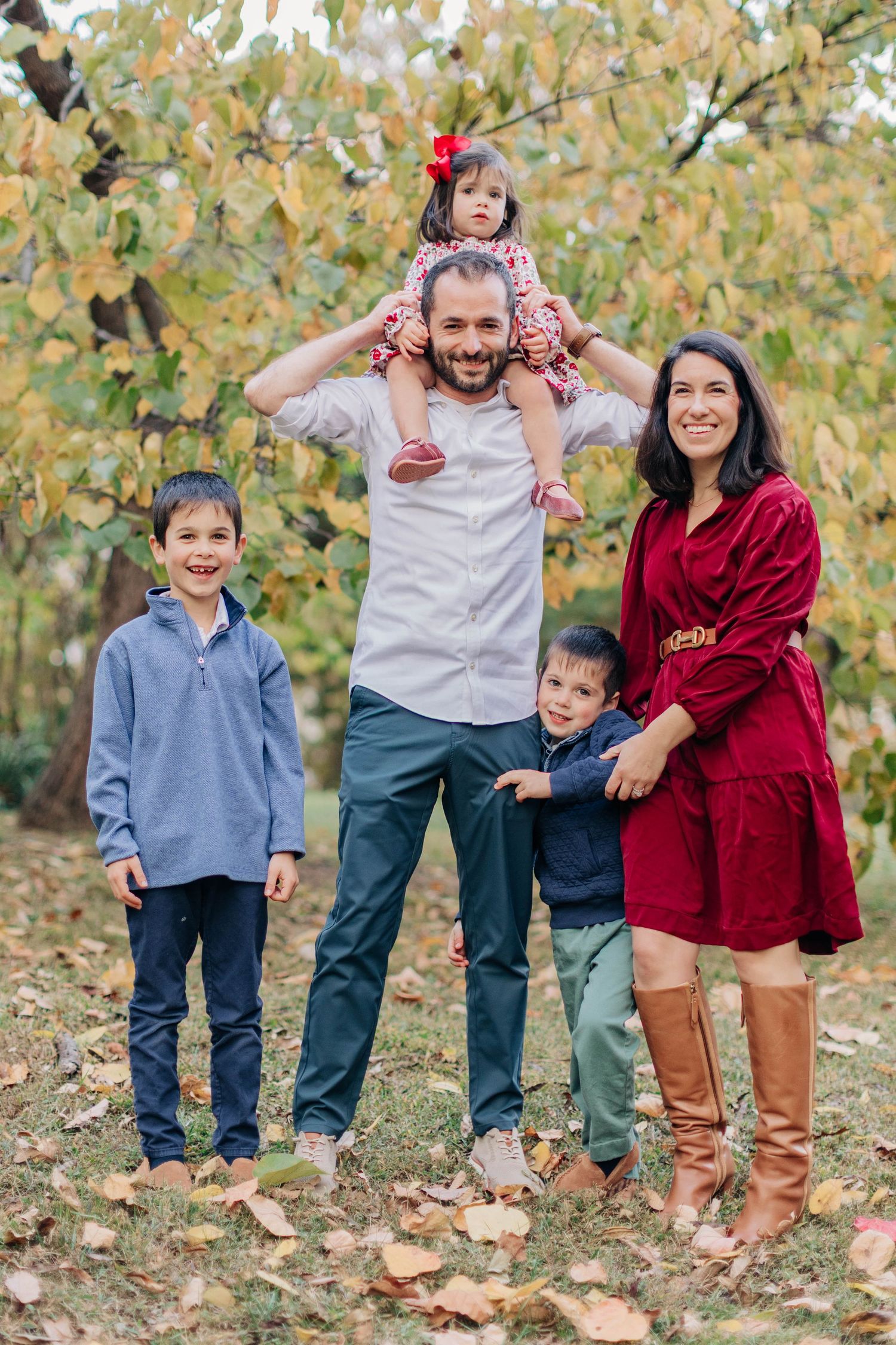 A happy family of five poses outdoors in autumn, with dad holding a toddler girl on his shoulders.