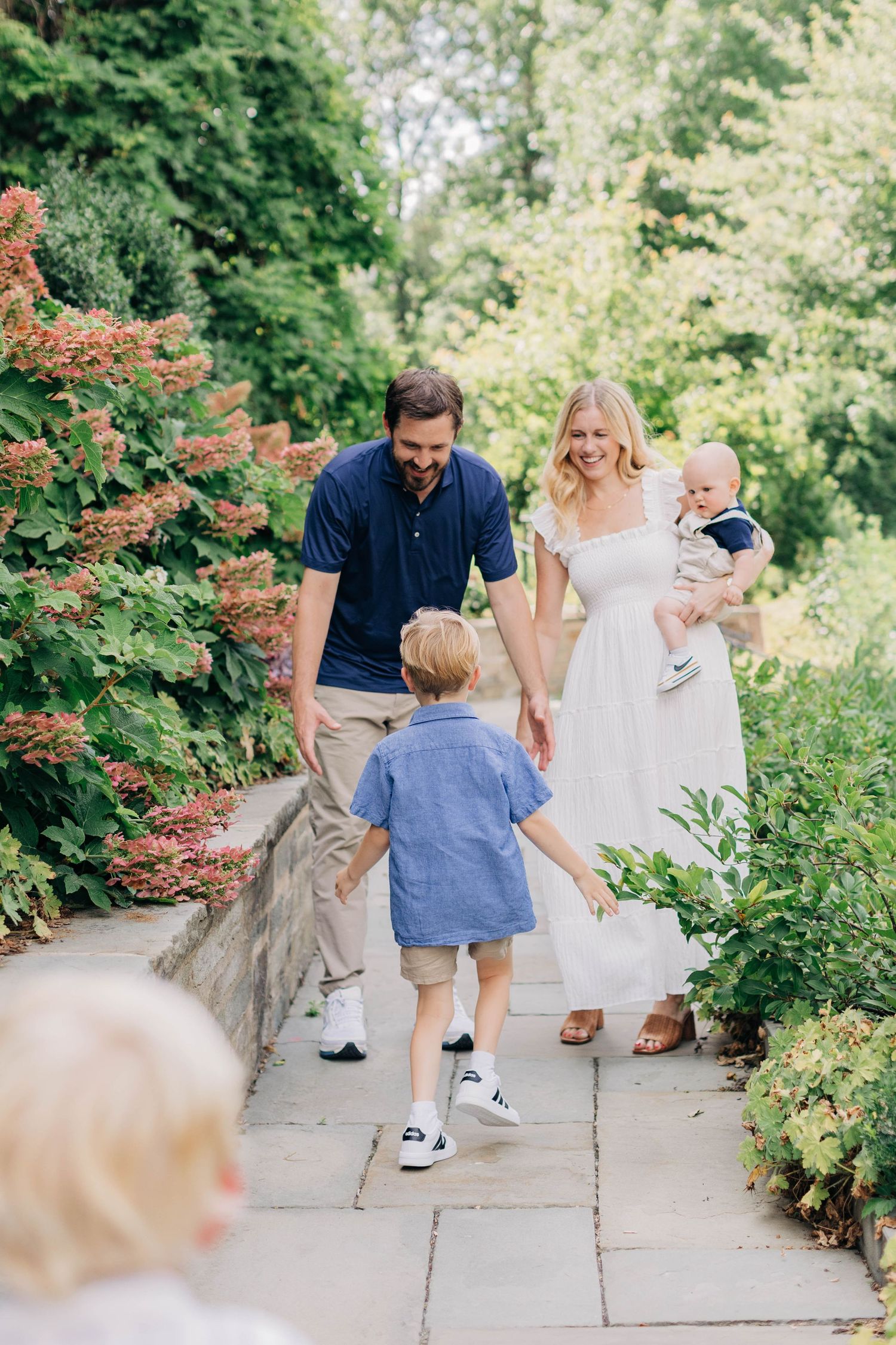 A happy family walks along a garden path, with parents and two young children surrounded by lush greenery.