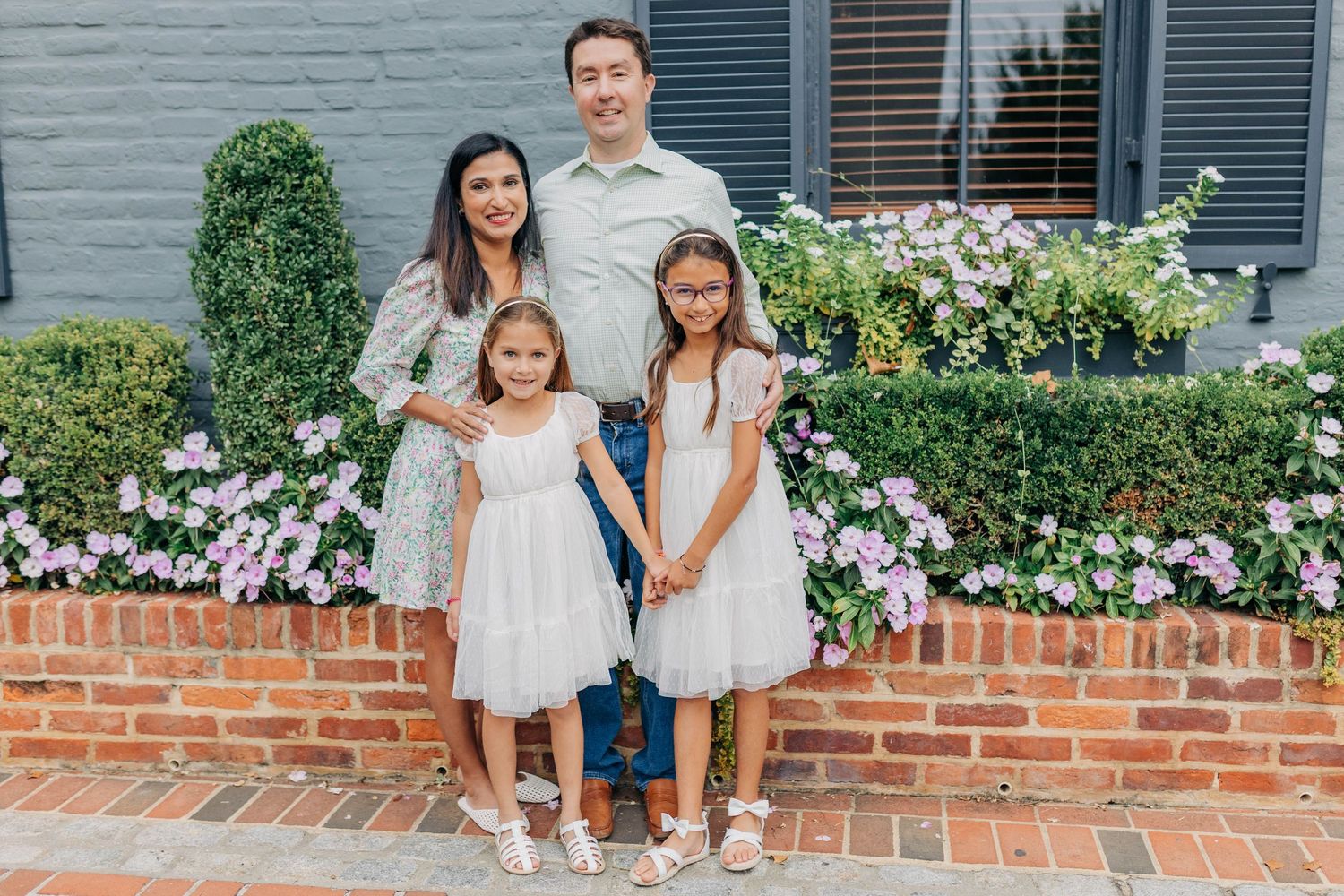 Family of four posing outdoors in white dresses near brick wall with pink flowers.