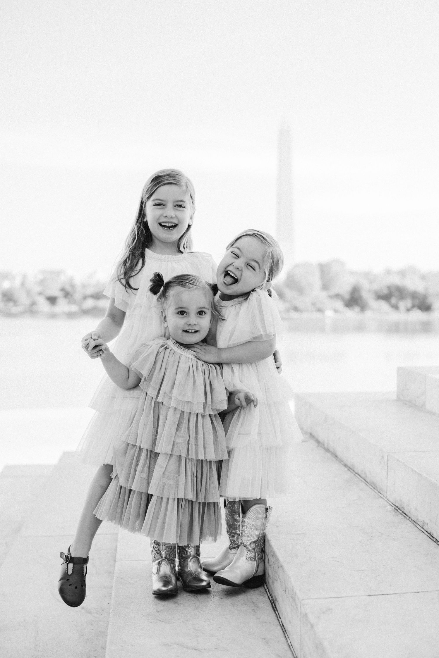 Three young girls in elegant dresses laugh and hug on marble steps near the Washington Monument in a black and white photo.