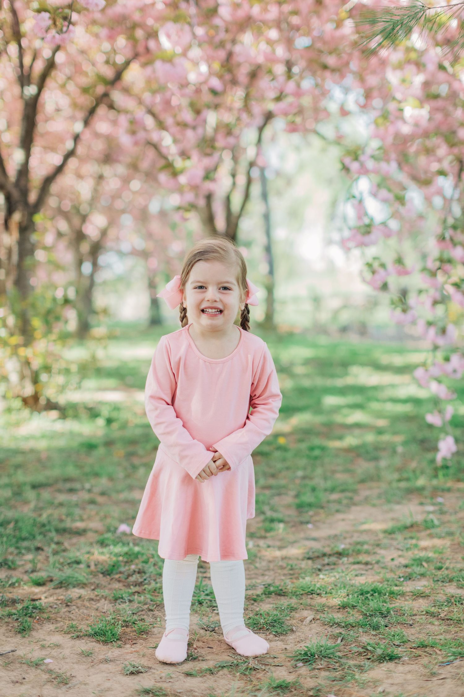 Young child in pink dress smiling while standing under cherry blossoms.