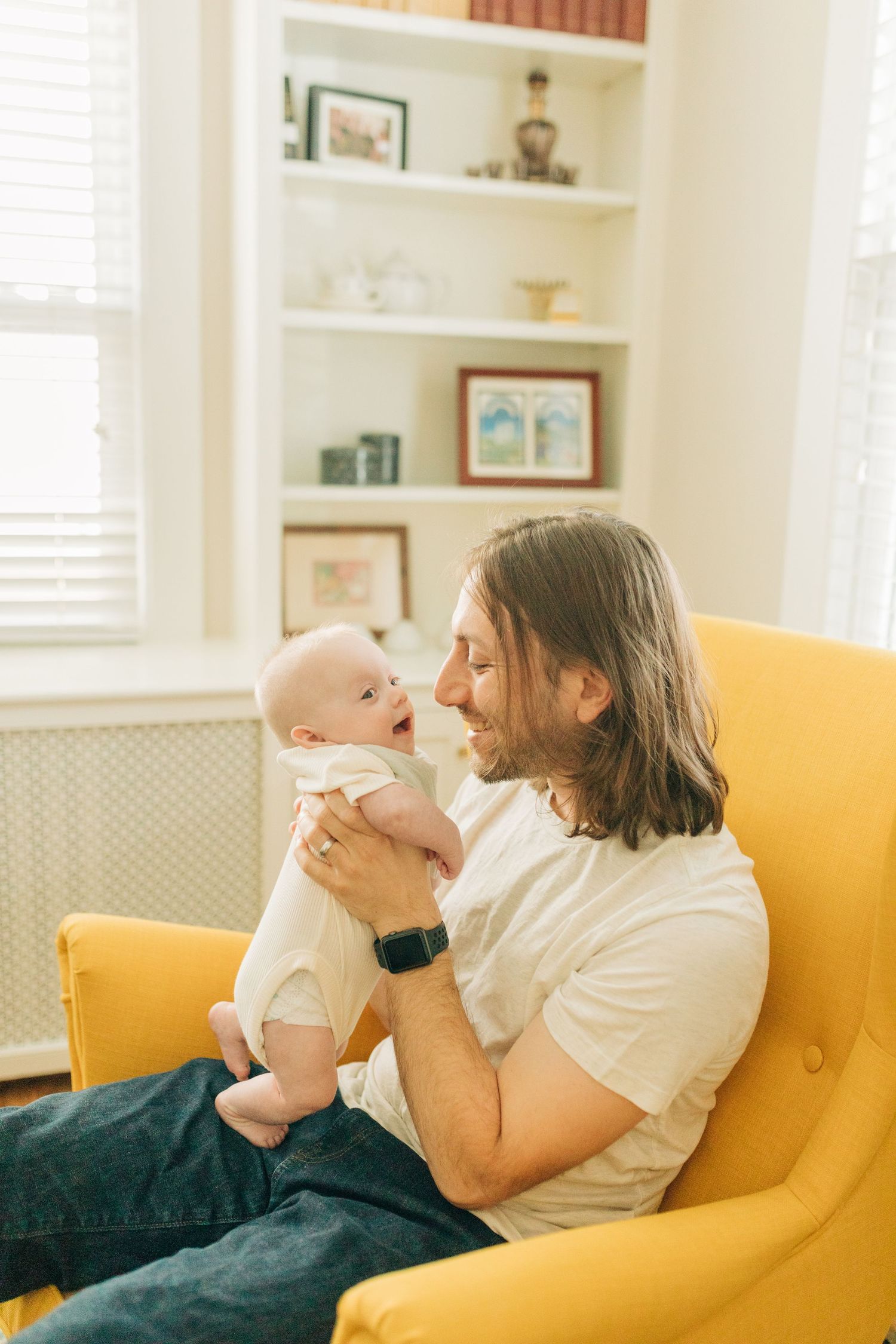 Tender moment shared on a vibrant yellow armchair in a sunlit room with built-in shelving.