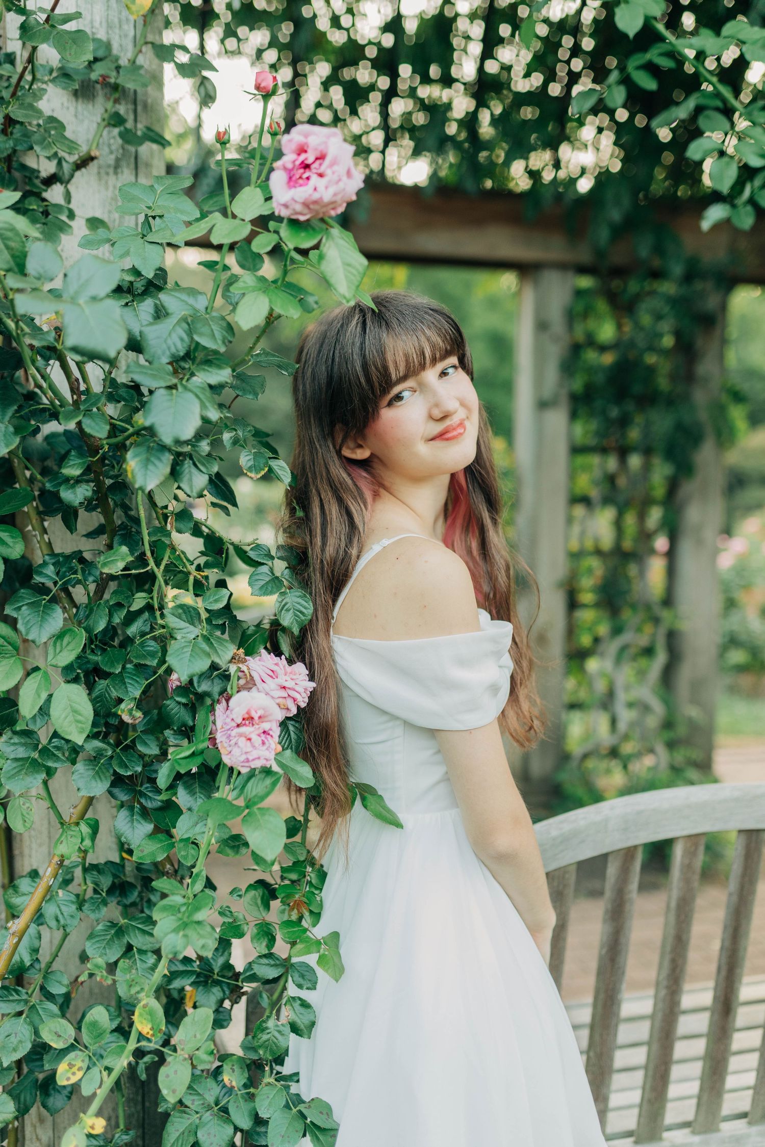 Someone in white dress poses by pink climbing roses on a garden trellis.
