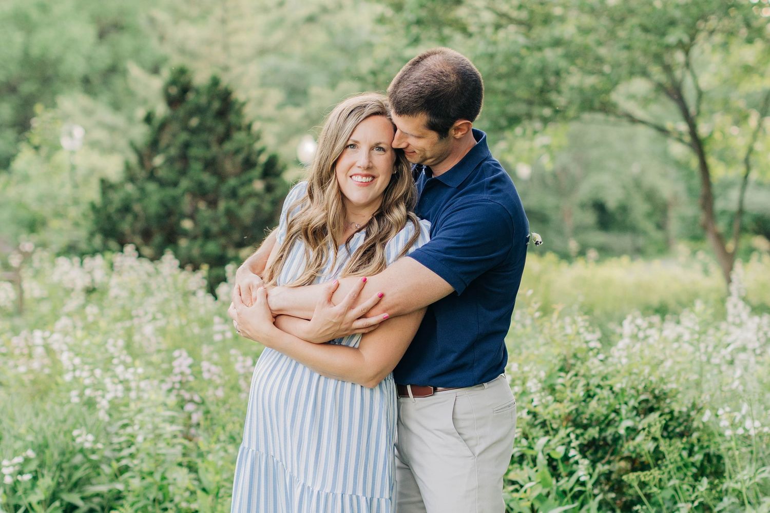 A couple embraces while standing in a sunlit garden with green foliage in the background.