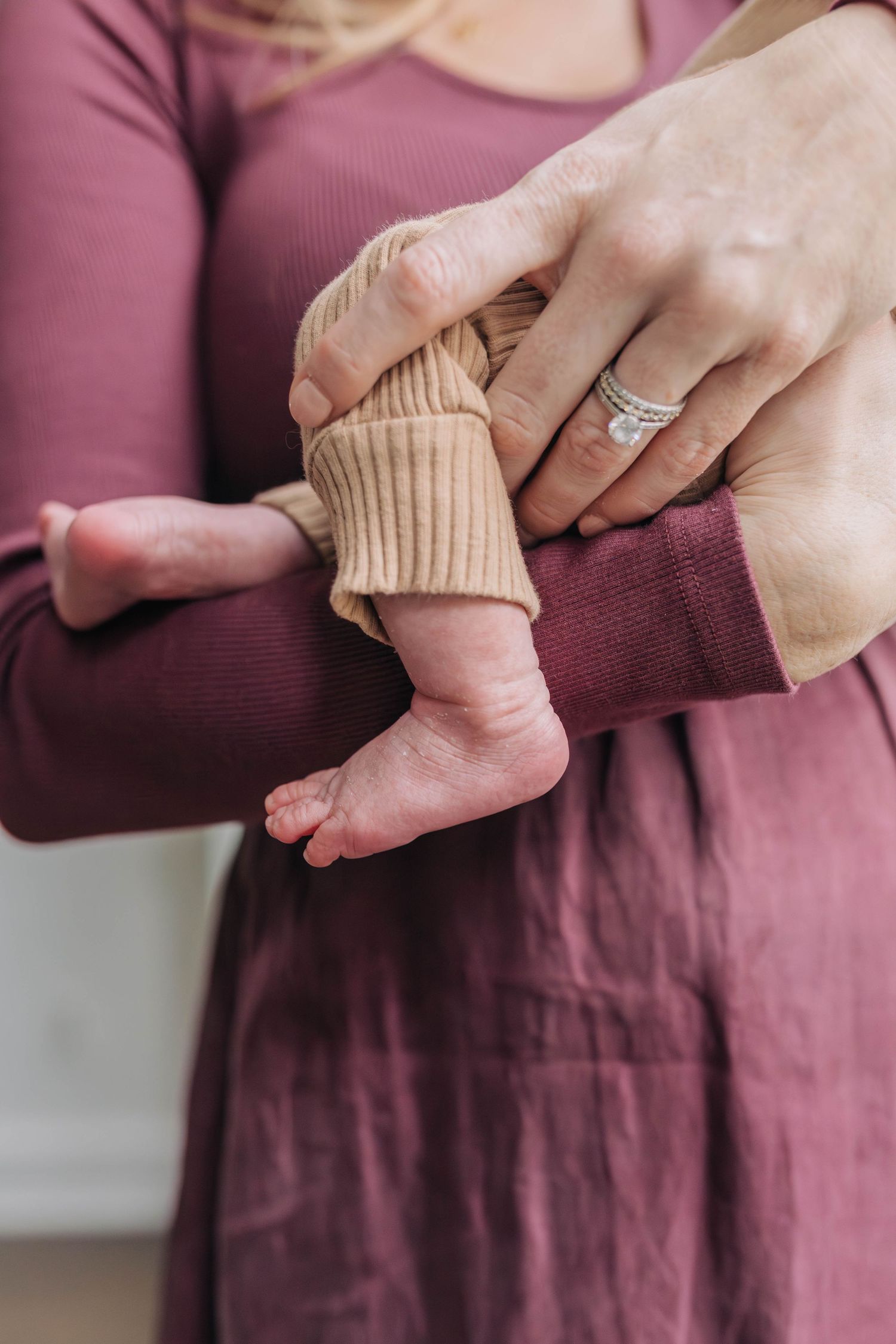 Close-up of tiny baby feet in beige knit clothing with mother's hands showing engagement ring.
