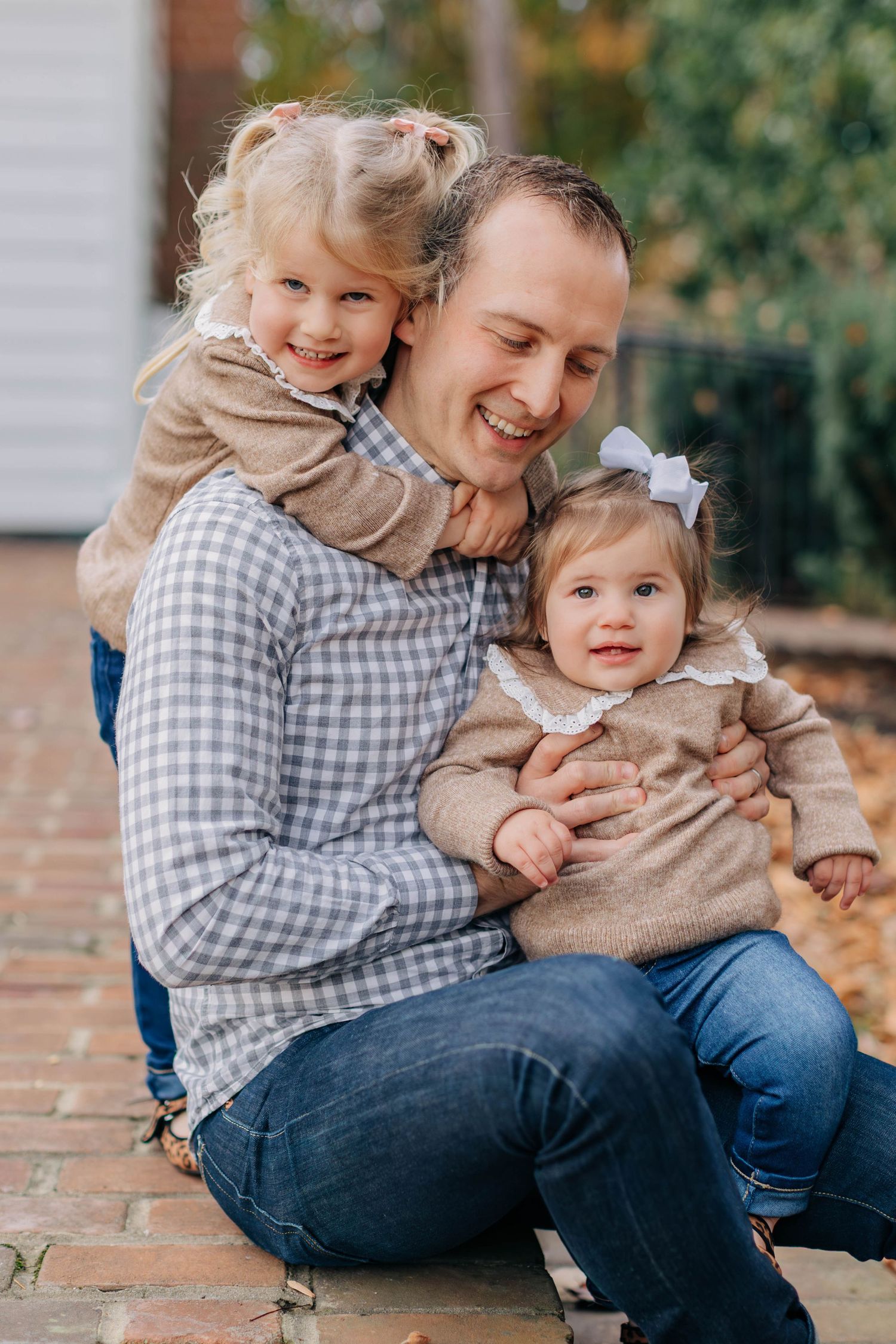A smiling father sits outdoors holding two young daughters in matching tan sweaters on a brick path.