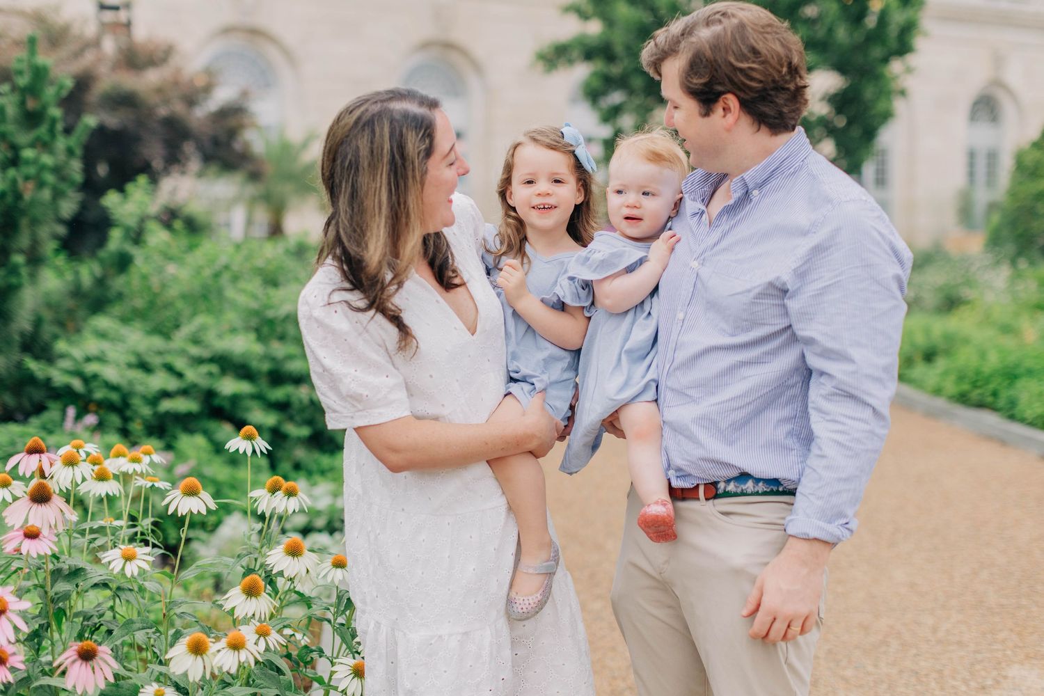 Family of four poses outdoors near blooming flowers, parents holding two young girls in matching blue dresses.