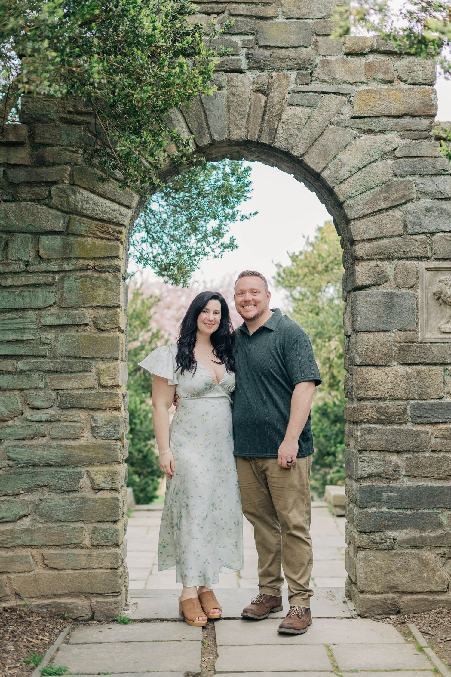 Smiling couple standing together under a rustic stone archway surrounded by lush greenery in an outdoor garden setting.