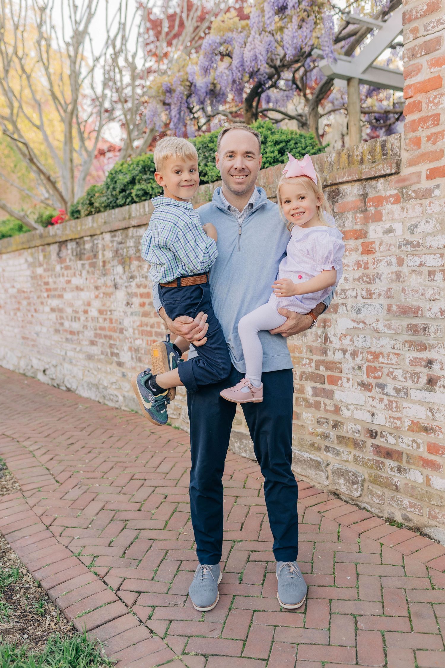 A smiling father holds his young son and daughter outdoors near a brick wall with purple wisteria blooms.