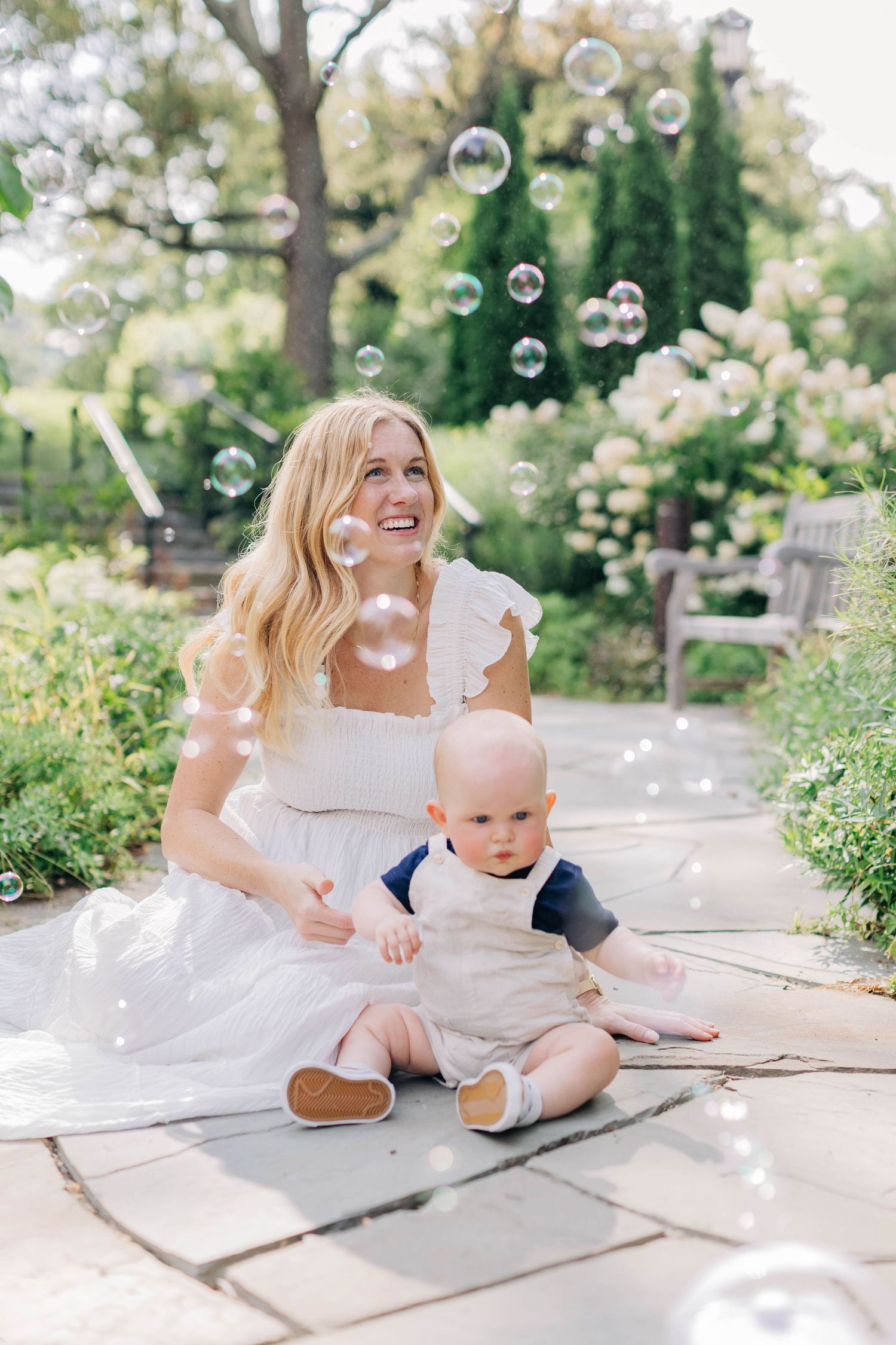 Smiling blonde mother in white dress sitting outdoors with baby surrounded by floating soap bubbles in garden.