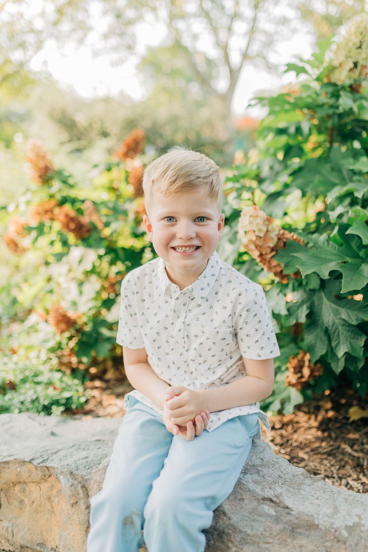 Child in polka dot shirt and jeans sits smiling on stone wall with greenery behind.