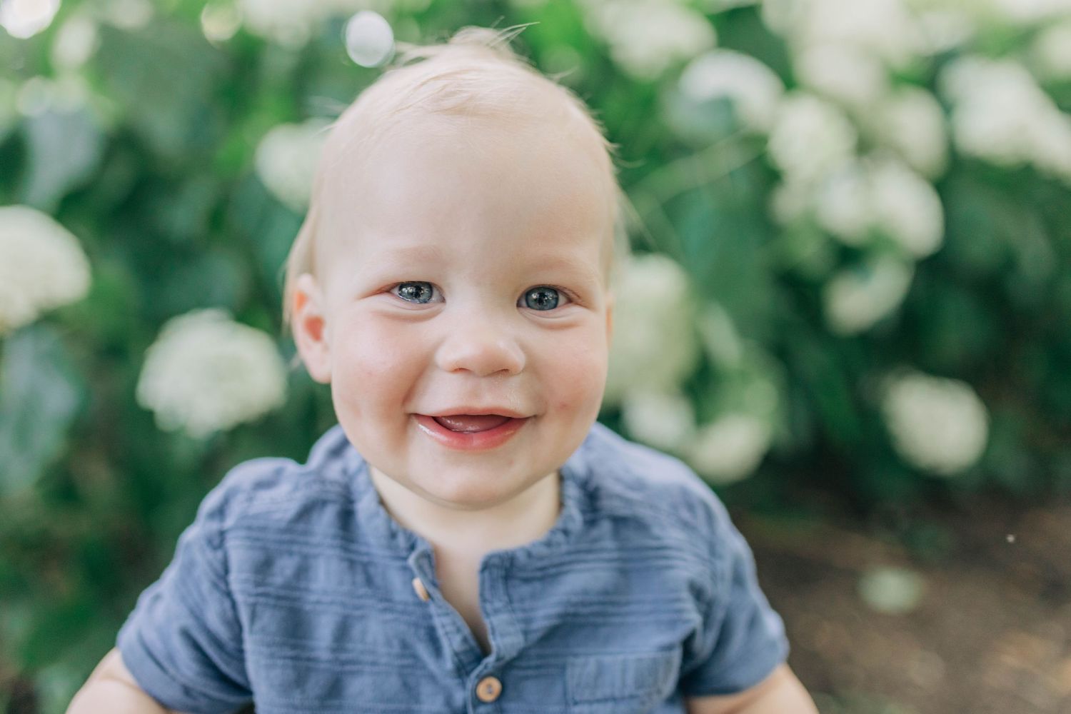 Baby in blue shirt and plaid shorts smiles while sitting outdoors with white flowers in background.