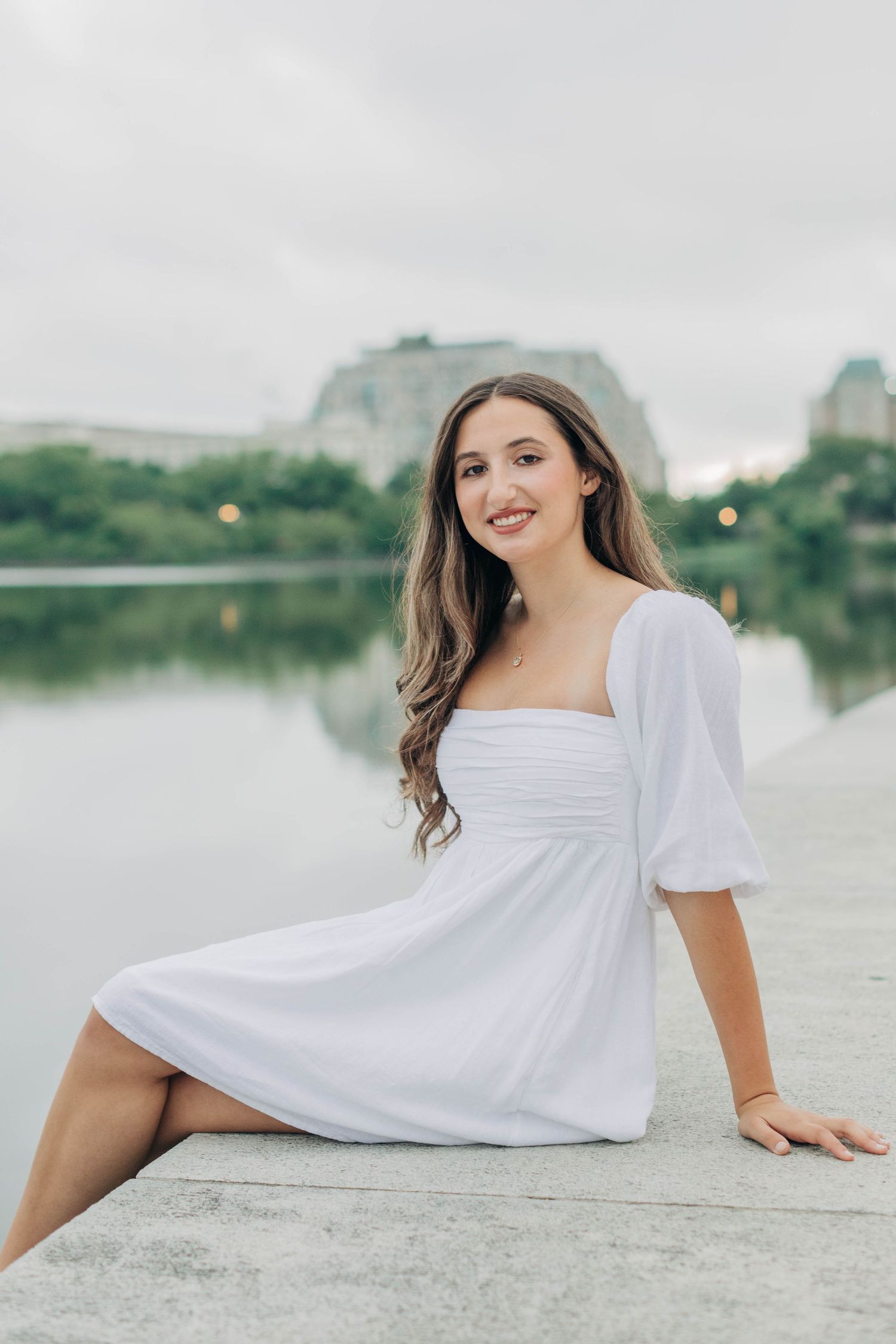 Person in a flowing white dress poses along a waterfront with cloudy skies in the background.