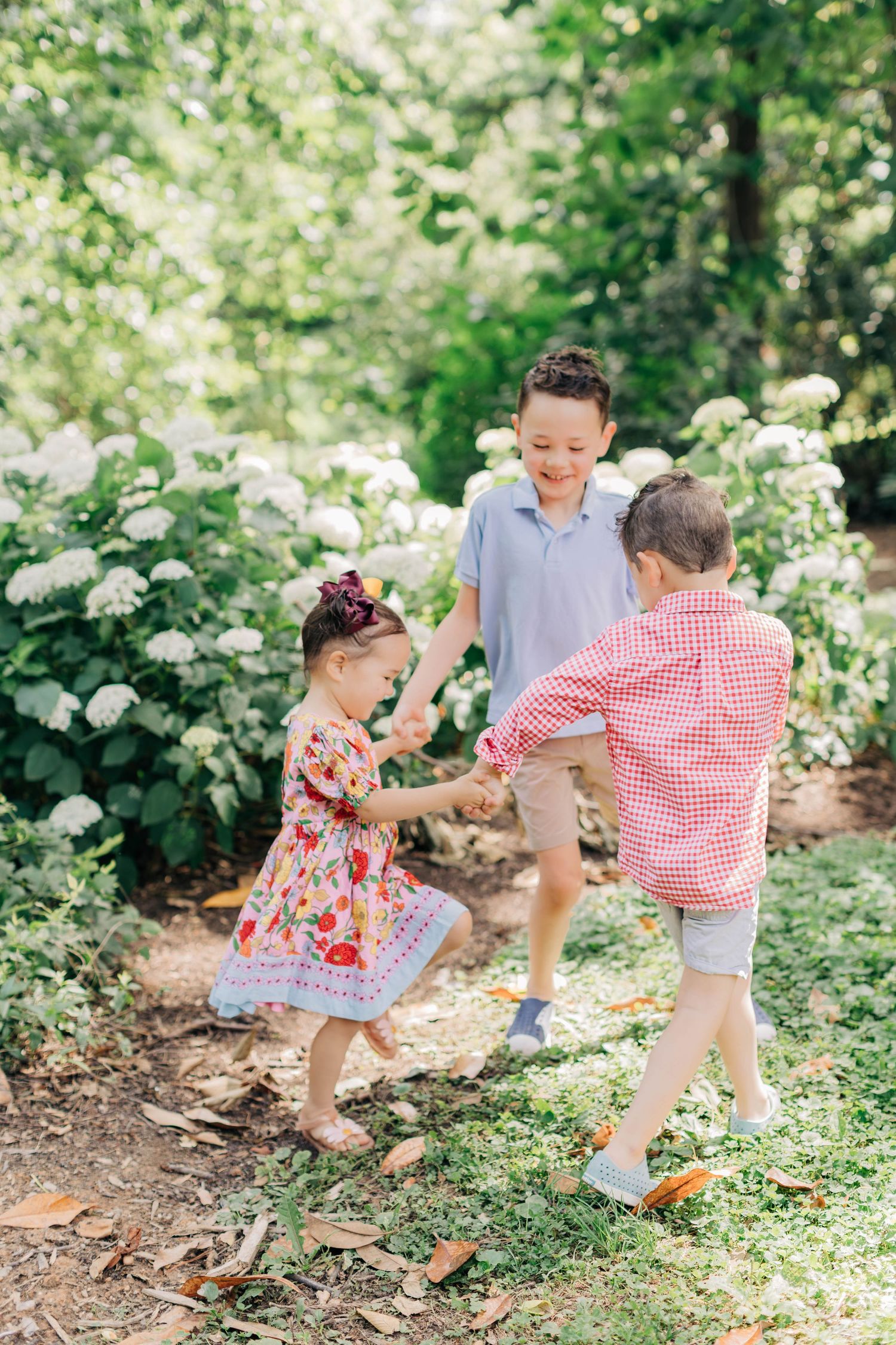 Three young children hold hands and play together in a lush garden with white hydrangea flowers blooming in the background.