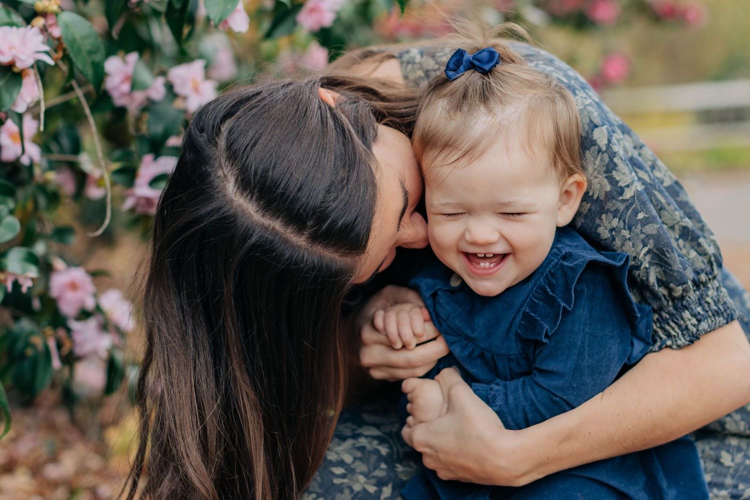 A mother tenderly kisses her laughing toddler girl in a blue dress near blooming pink flowers outdoors.