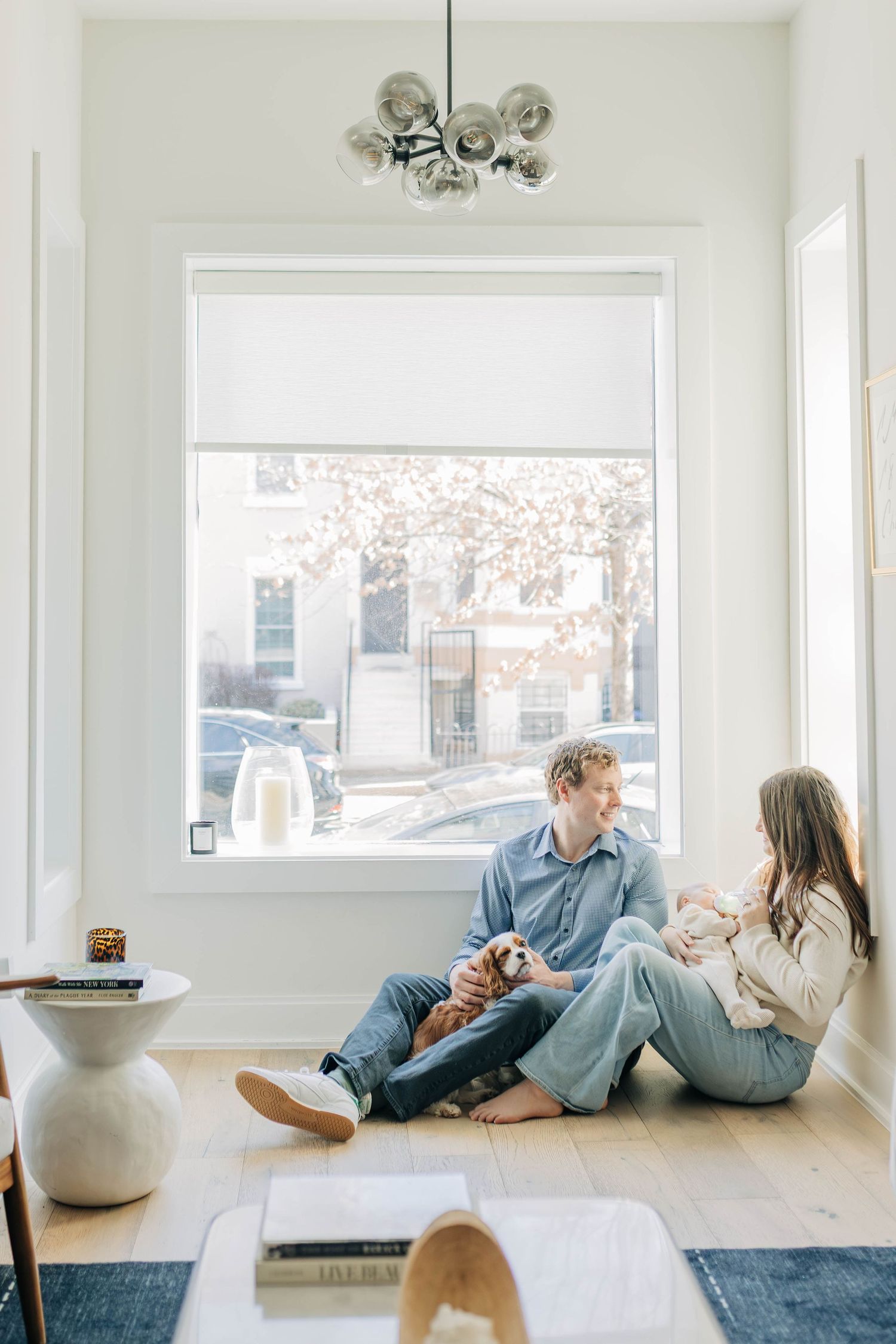 Young couple sitting on hardwood floor by large window with newborn baby and small dog in modern home.