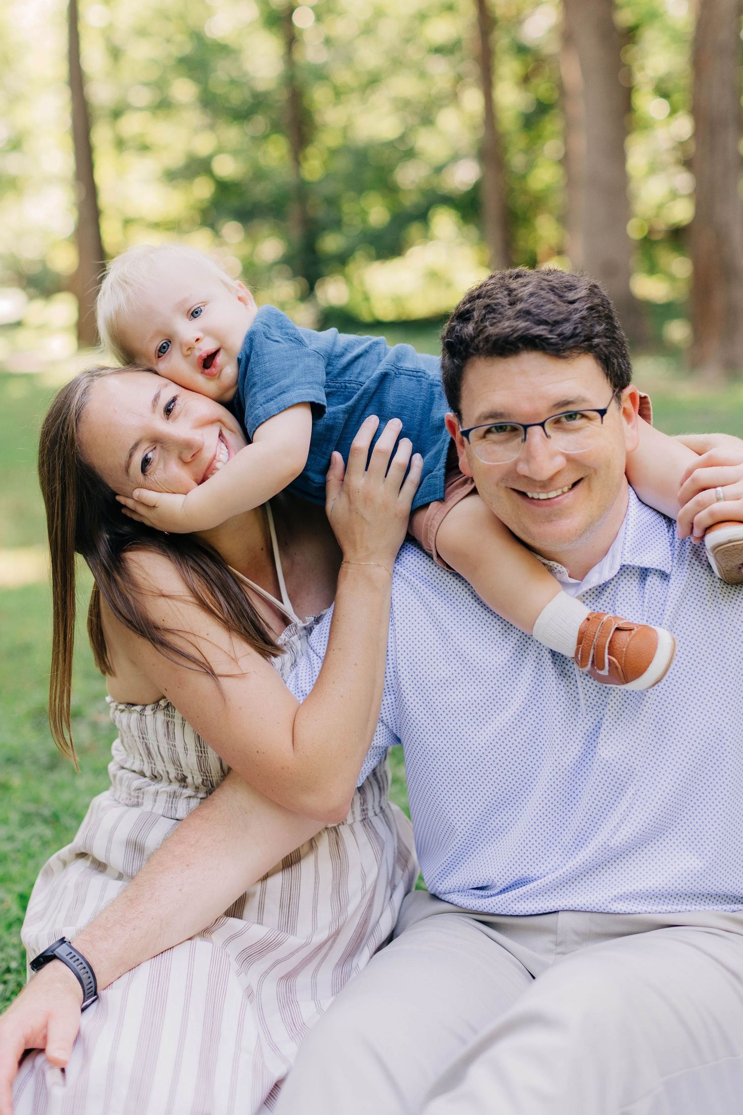 Happy young family with baby boy sitting outdoors in green park, smiling together in casual summer attire.