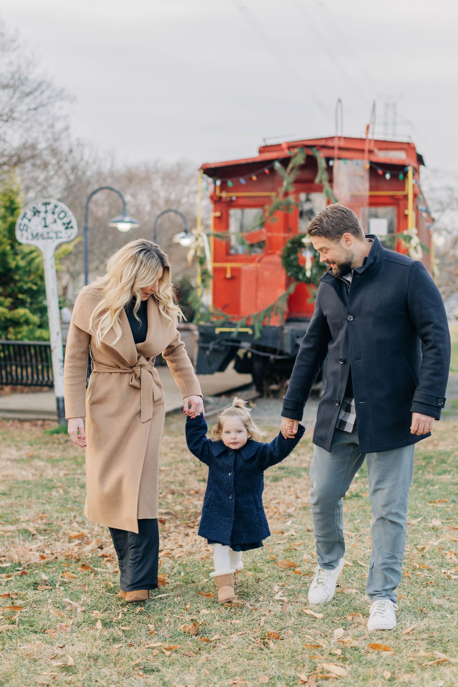 Family of three walking outdoors in winter coats, holding toddler's hands near a decorated red train car.